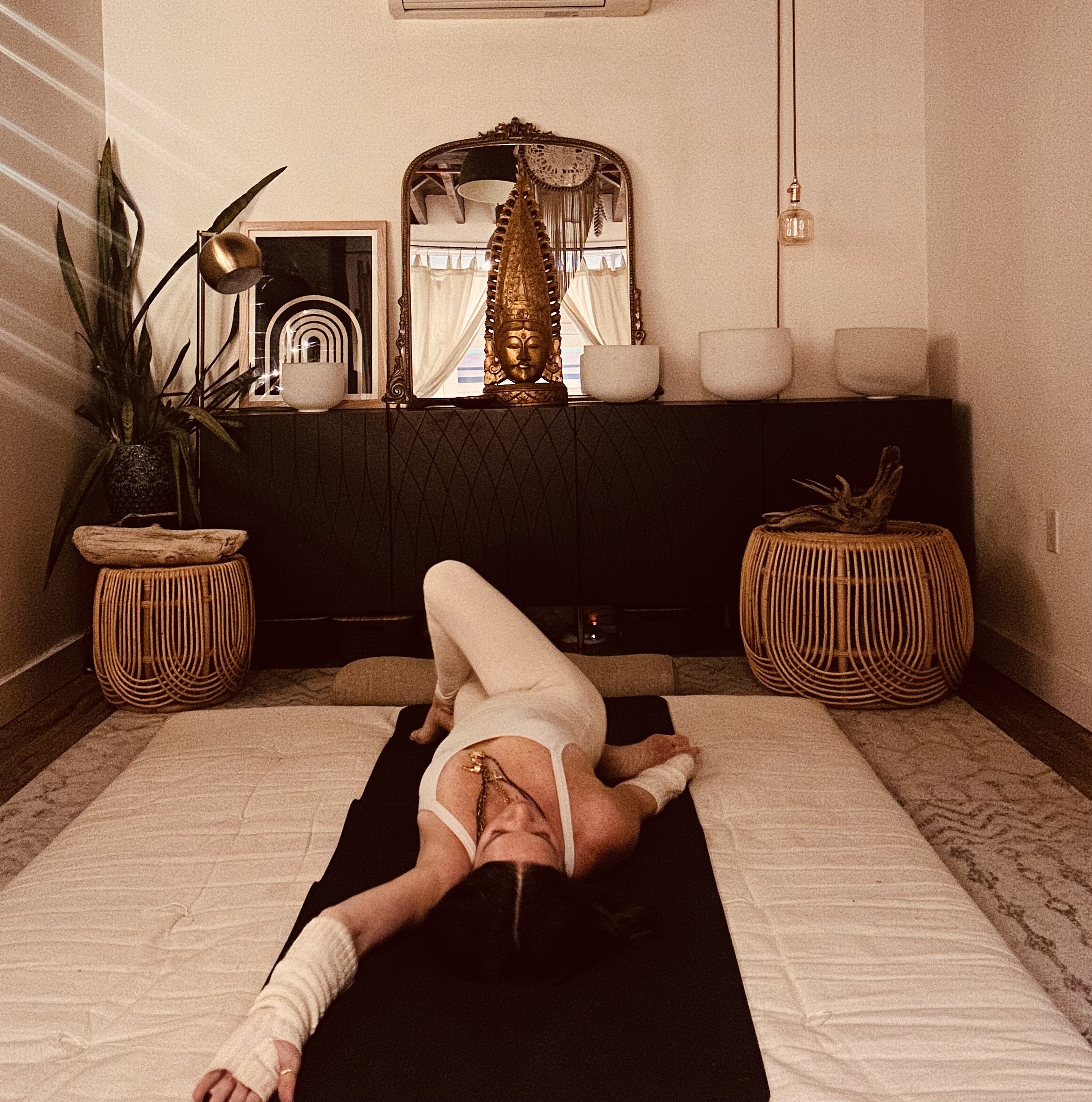 A woman practicing yoga or meditation on a mat on the floor in a cozy, dimly lit room, with decorative items and plants in the background.