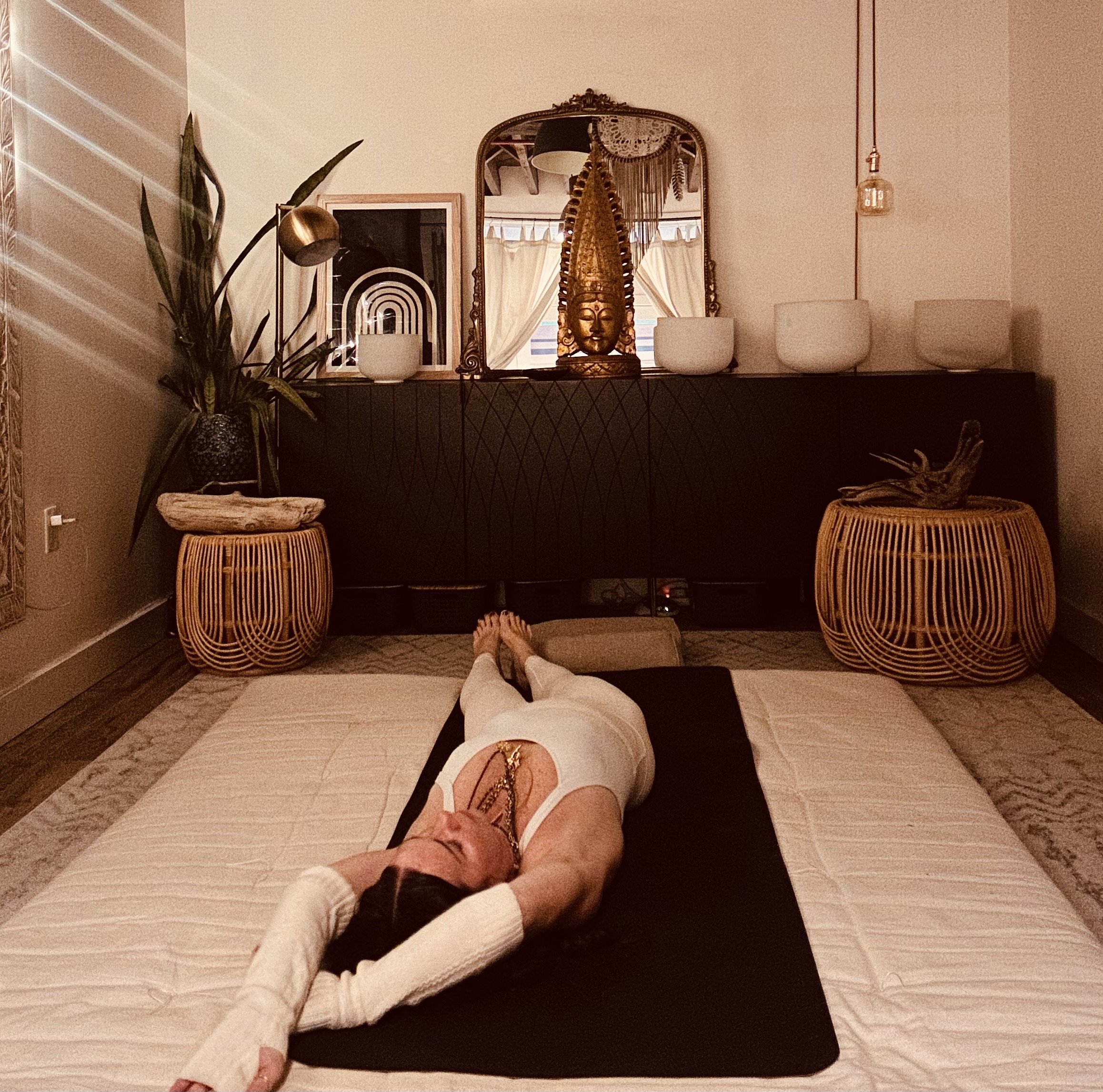 A woman lying on her back on a black yoga mat on a bed with her hands behind her head, in a room with warm lighting, a mirror, decorative statues, vases, and plants.