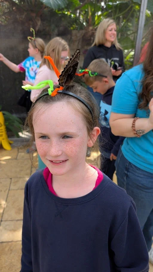 A young girl with a butterfly headband, standing outdoors with other children and an adult in the background.