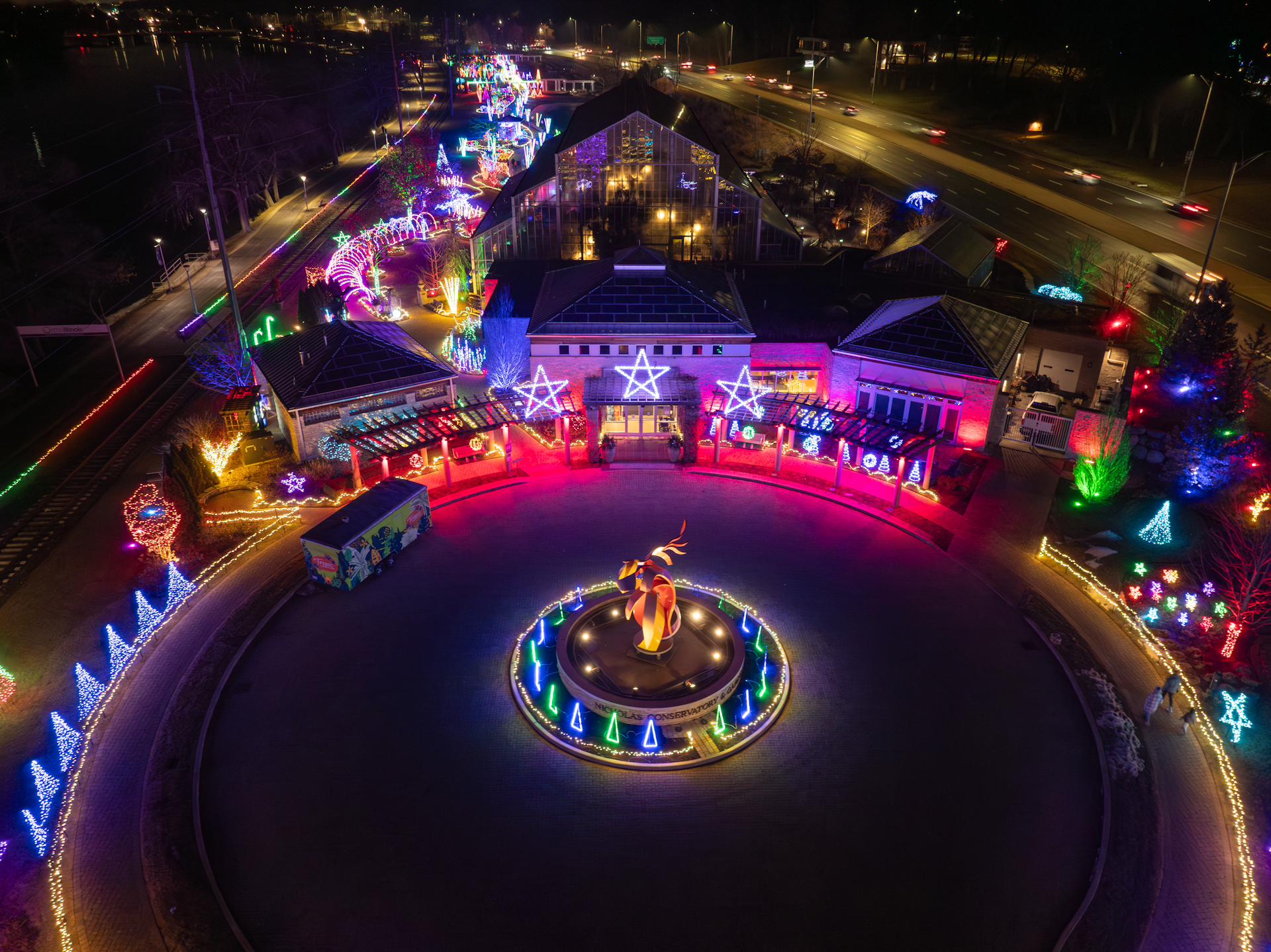Nighttime scene of a park decorated with colorful Christmas lights, including arches, trees, star-shaped ornaments, and a cone-shaped structure.
