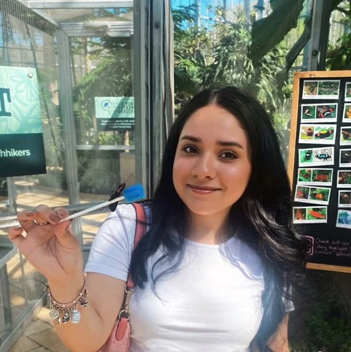 Young woman with dark hair smiling inside a greenhouse holding a blue toothbrush.