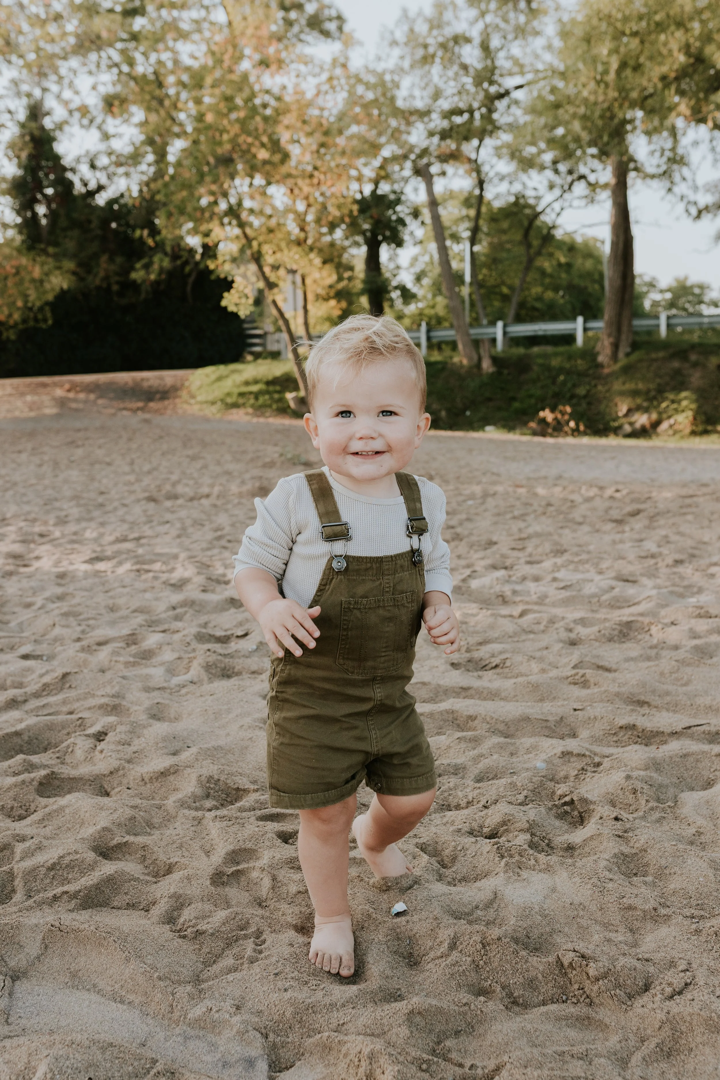baby boy walking barefoot and smiling for photos during a mini photo session at de la salle beach in georgina ontario
