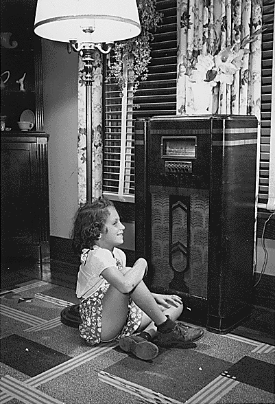 Image: Black and White photo of 1930's house setting, smiling young girl sitting cross-legged in front of a standing radio. 