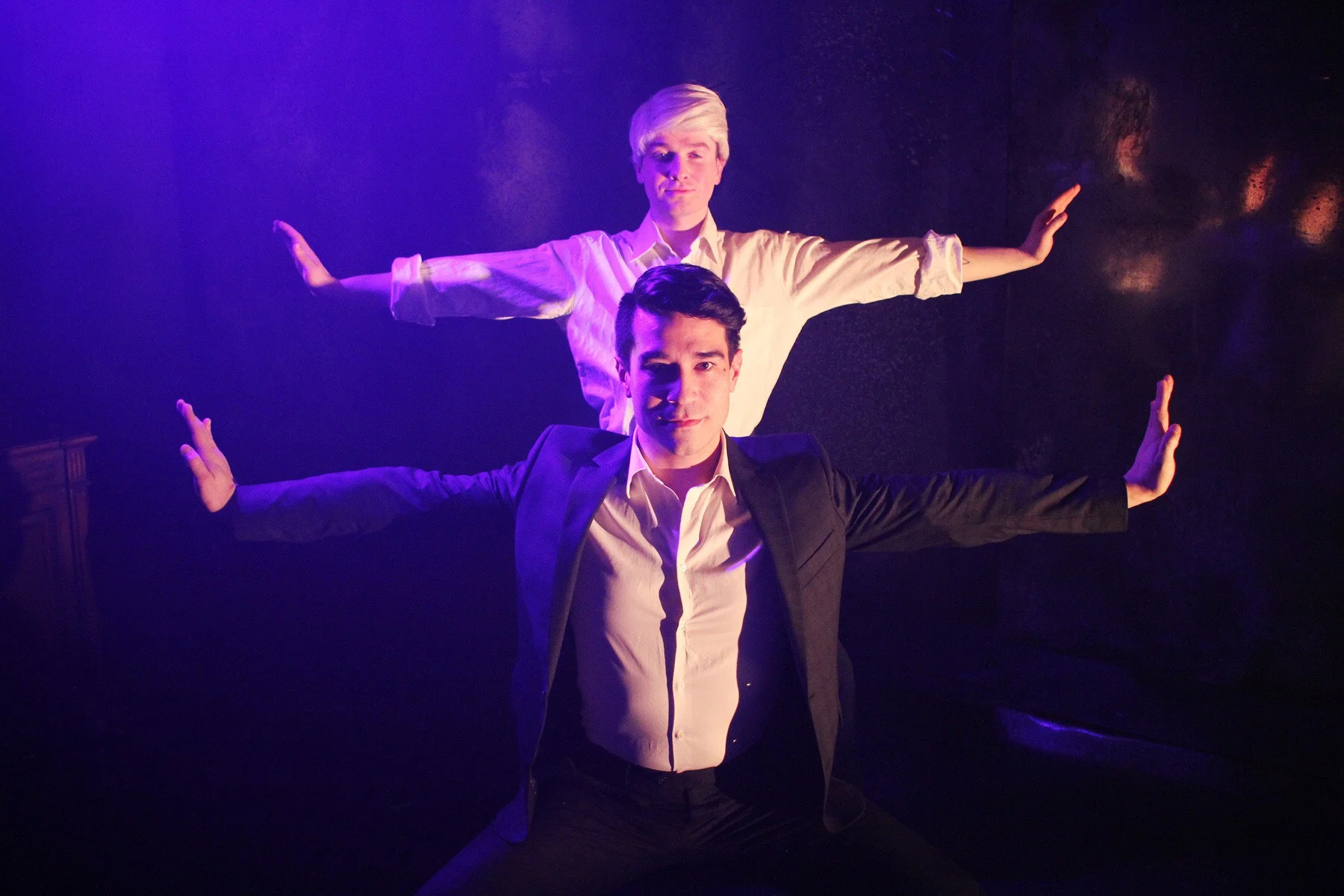 Two men in formal attire are standing with their arms extended outward, one in front and one behind, against a dark background with purple lighting.