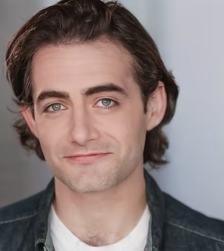 A young man with brown hair, blue eyes, and fair skin smiling softly, wearing a dark denim jacket and a white shirt.