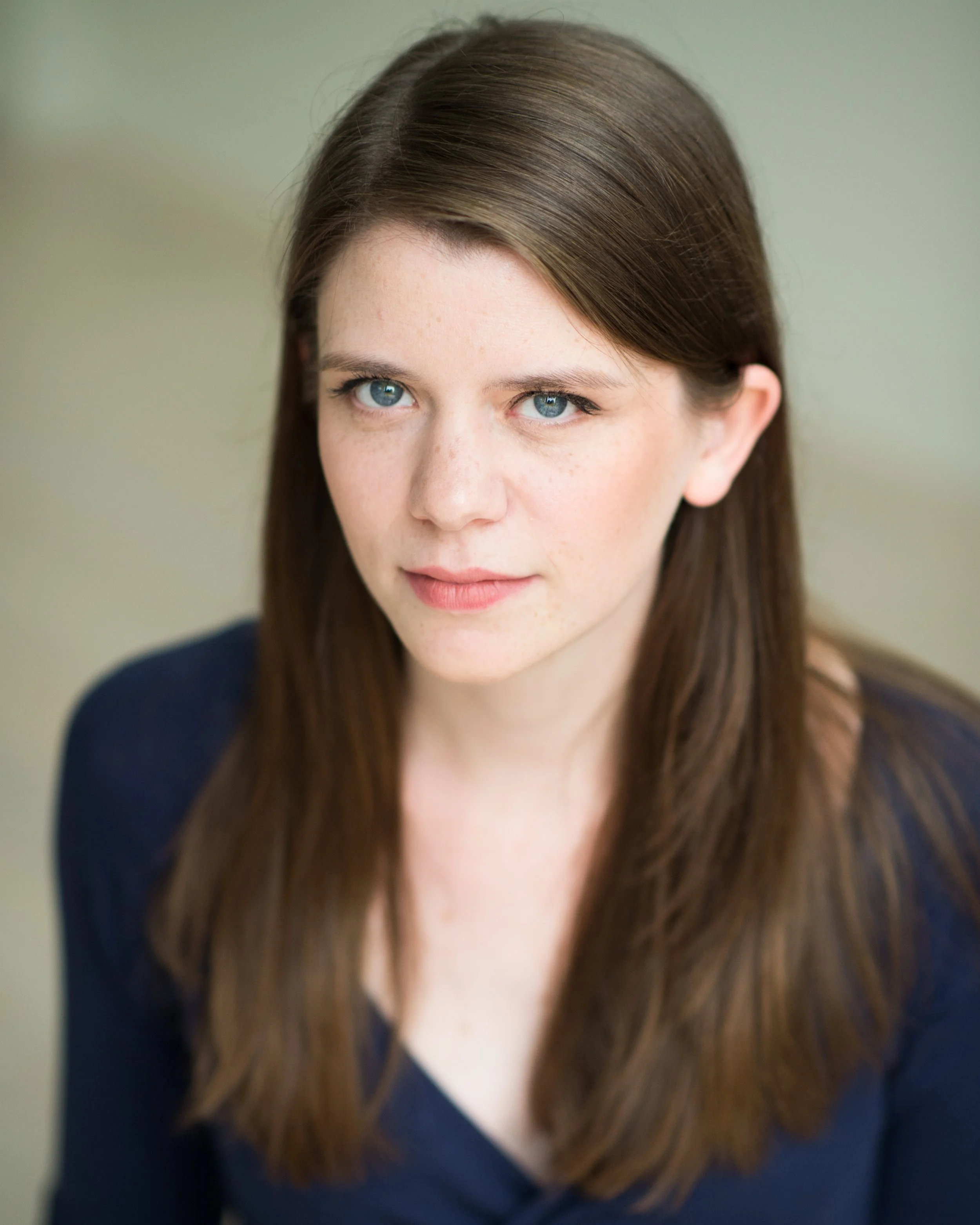 Close-up of a woman with straight, brown hair and blue eyes, wearing a dark top, looking into the camera.