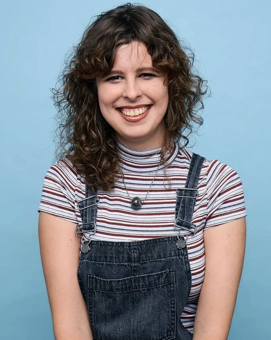 A young woman with curly brown hair, smiling and winking at the camera, wearing a striped t-shirt, denim overalls, and a necklace, against a light blue background.