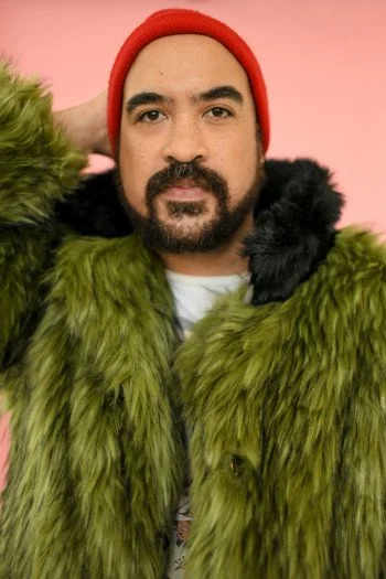 A man with a beard and mustache wearing a red beanie, green fur coat, and black and green accents, posing against a pink background.