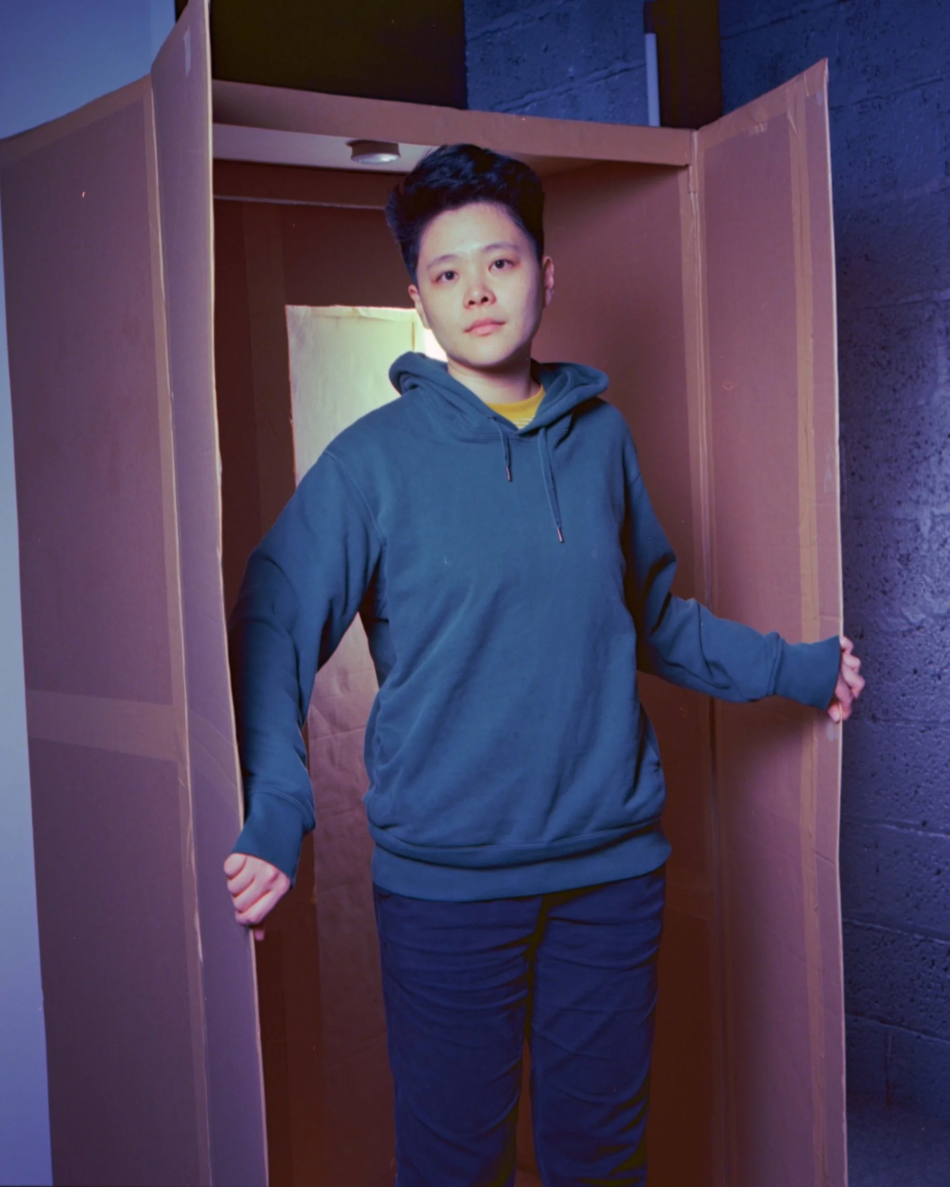 A person standing in a doorway, wearing a dark hoodie and pants, with one arm extended holding the door open. The person has short dark hair and a neutral facial expression.