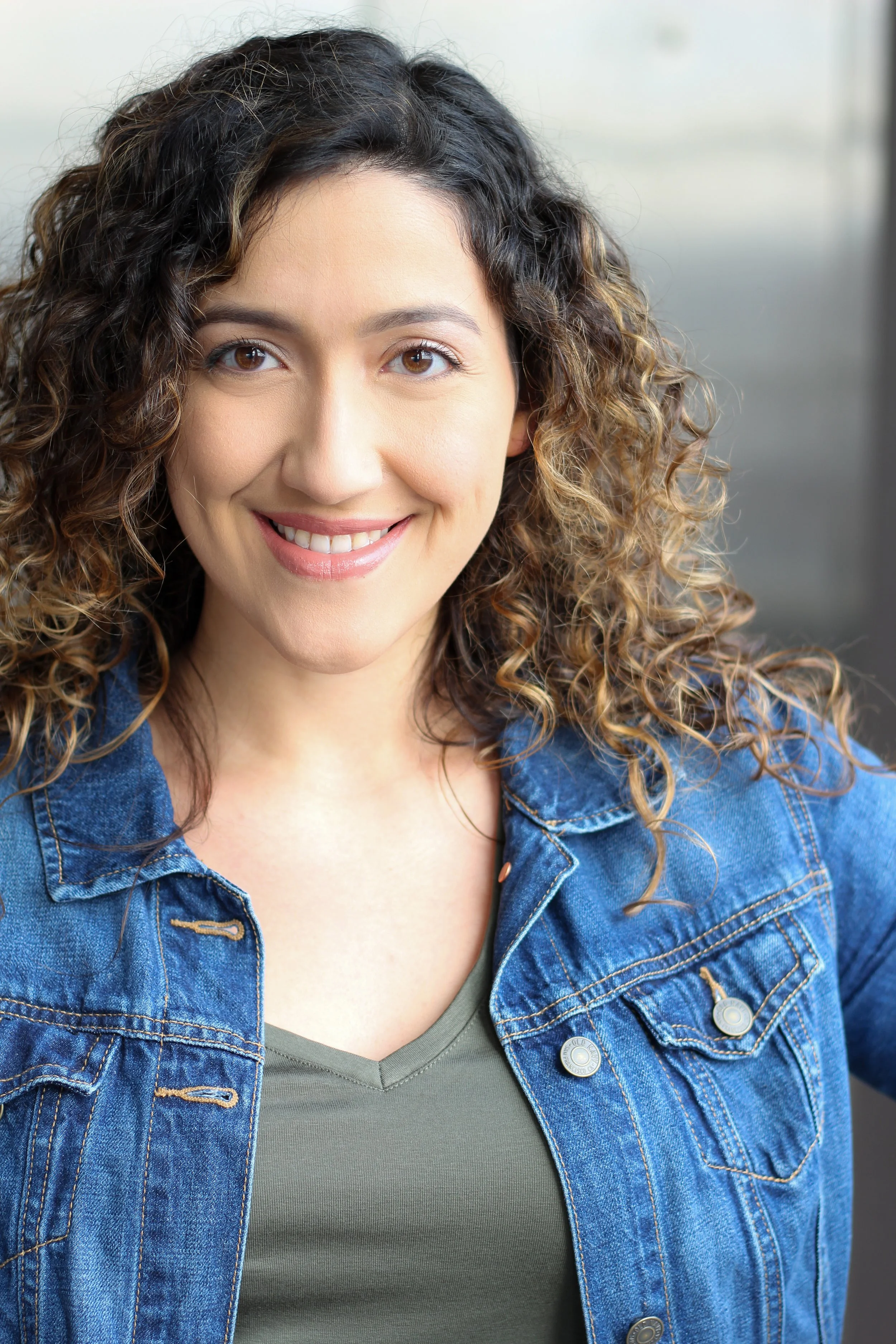 Close-up of a smiling woman with curly brown hair, wearing a blue denim jacket over a gray shirt, in front of a neutral background.