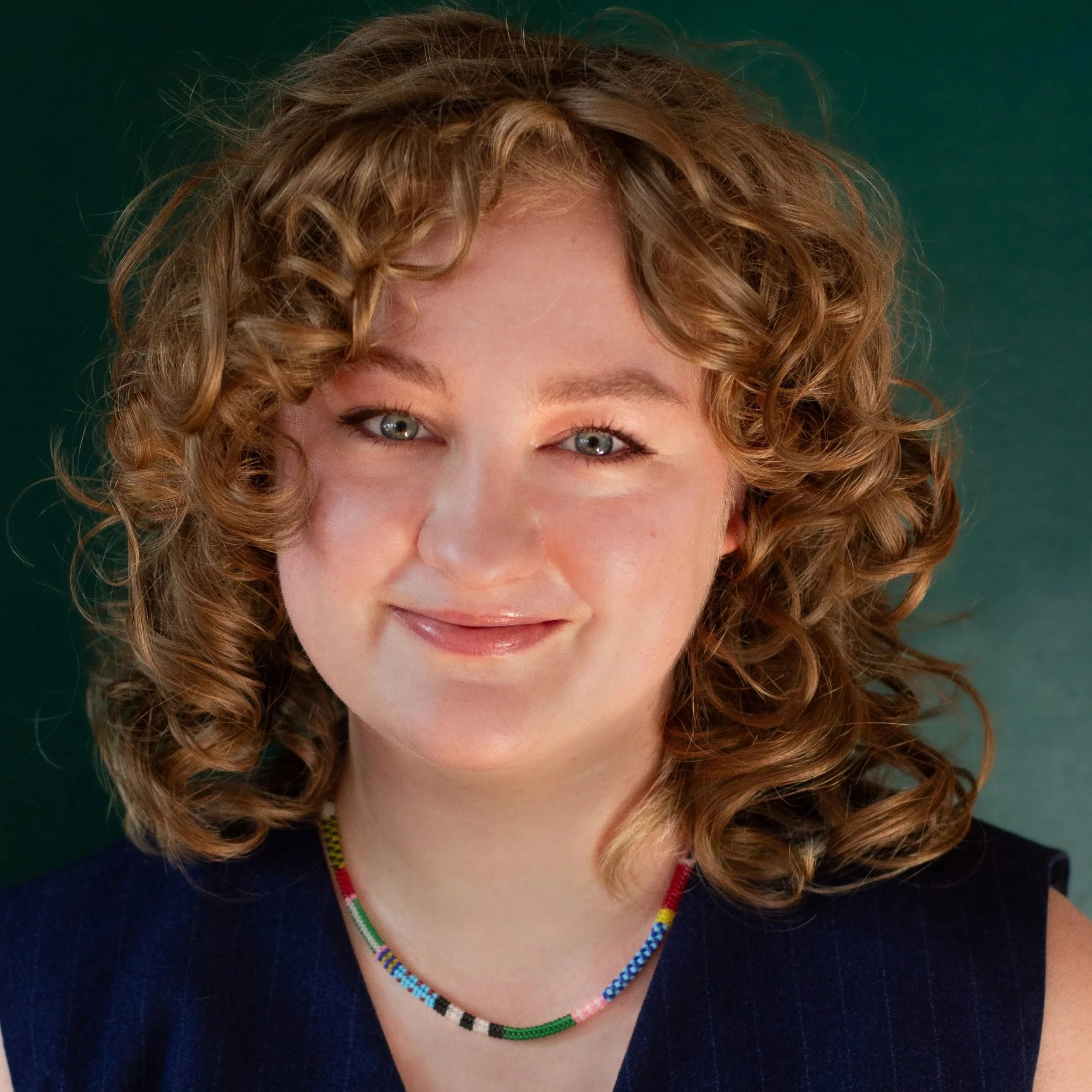 Close-up of a woman with curly reddish hair, wearing a colorful beaded necklace, smiling slightly, with a blurred teal background.