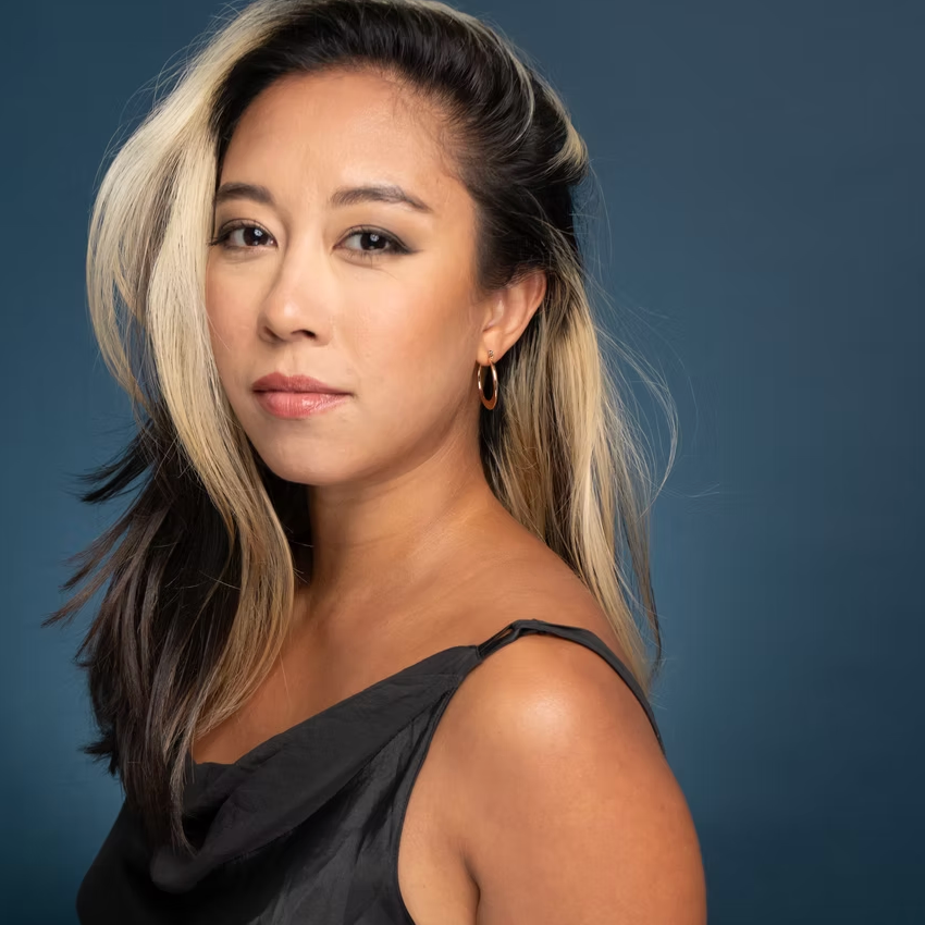 Close-up portrait of a woman with blonde and dark hair, wearing hoop earrings and a black sleeveless top, against a dark teal background.