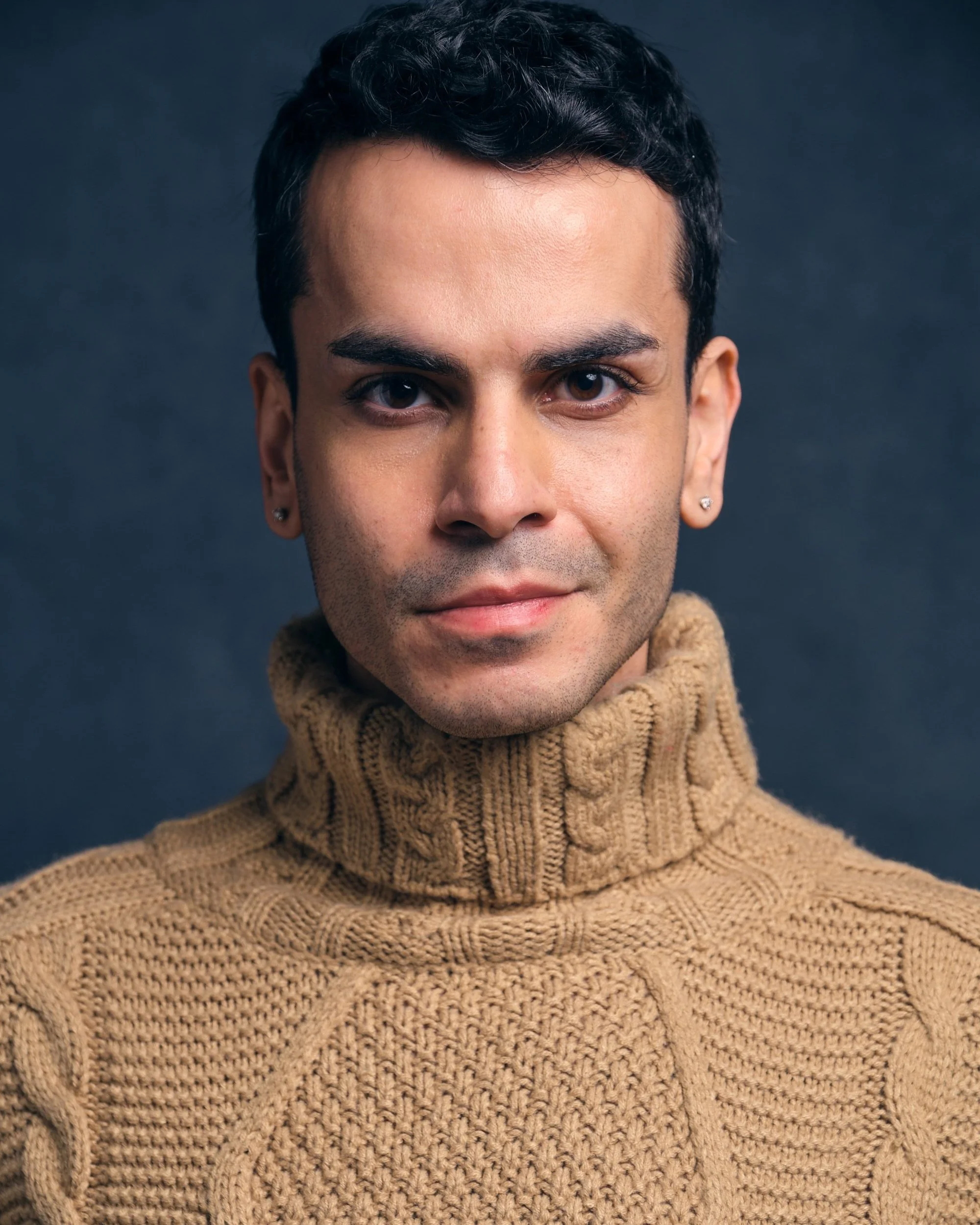 Close-up of a man with short black curly hair, wearing earrings and a brown knitted turtleneck sweater against a dark background.
