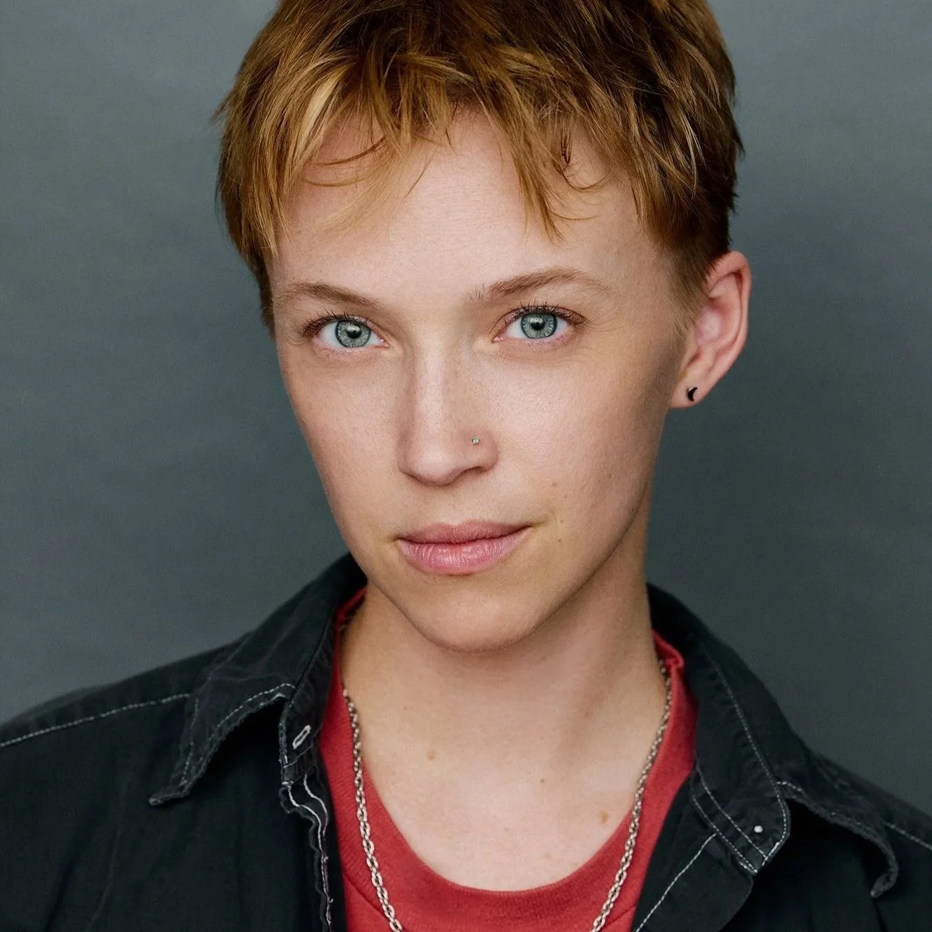 A young person with short, reddish hair and striking blue eyes looking directly at the camera. They have a nose piercing and are wearing black earrings, a red shirt, layered necklaces, and a dark button-up shirt over the red shirt. The background is a plain, neutral gray.