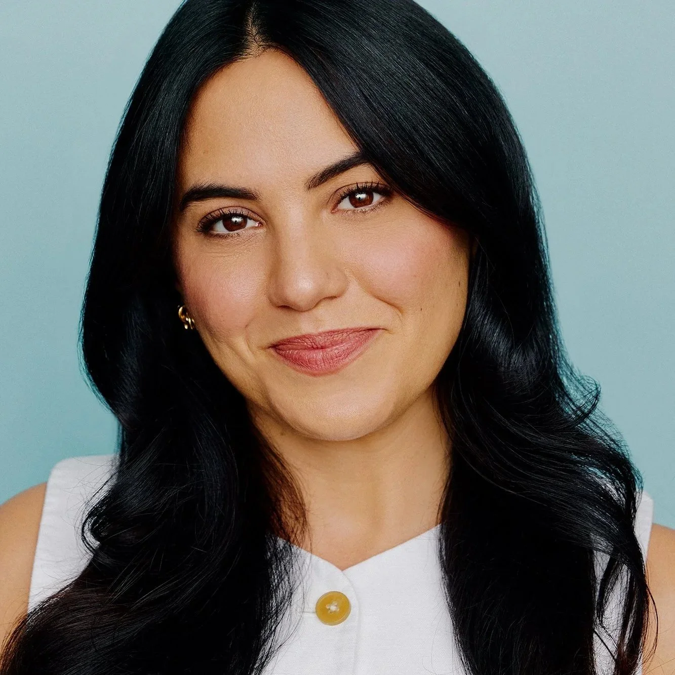 Close-up portrait of a woman with long black hair, wearing a white top with a yellow button, smiling slightly, against a light blue background.