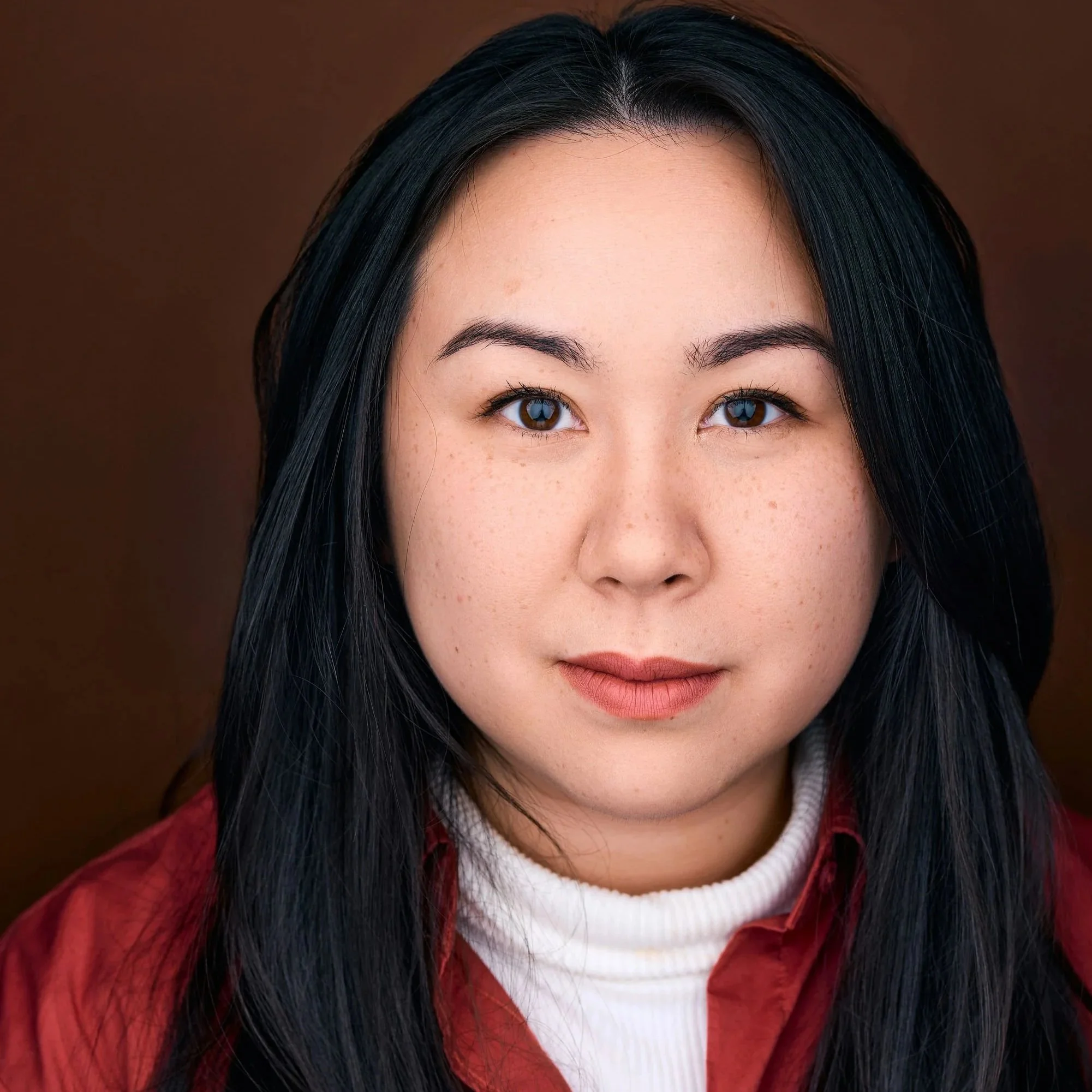 Close-up of a woman's face showing dark hair, brown eyes, and pink lips, with a blurred brown background.