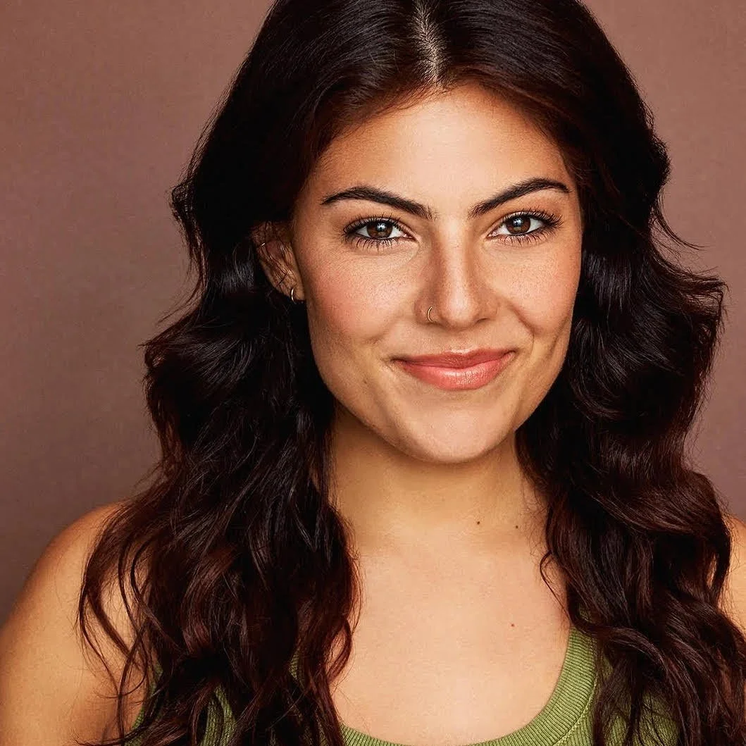 Close-up portrait of a young woman with long, wavy brown hair, fair skin, hazel eyes, and a subtle smile, wearing a green tank top against a plain brown background.