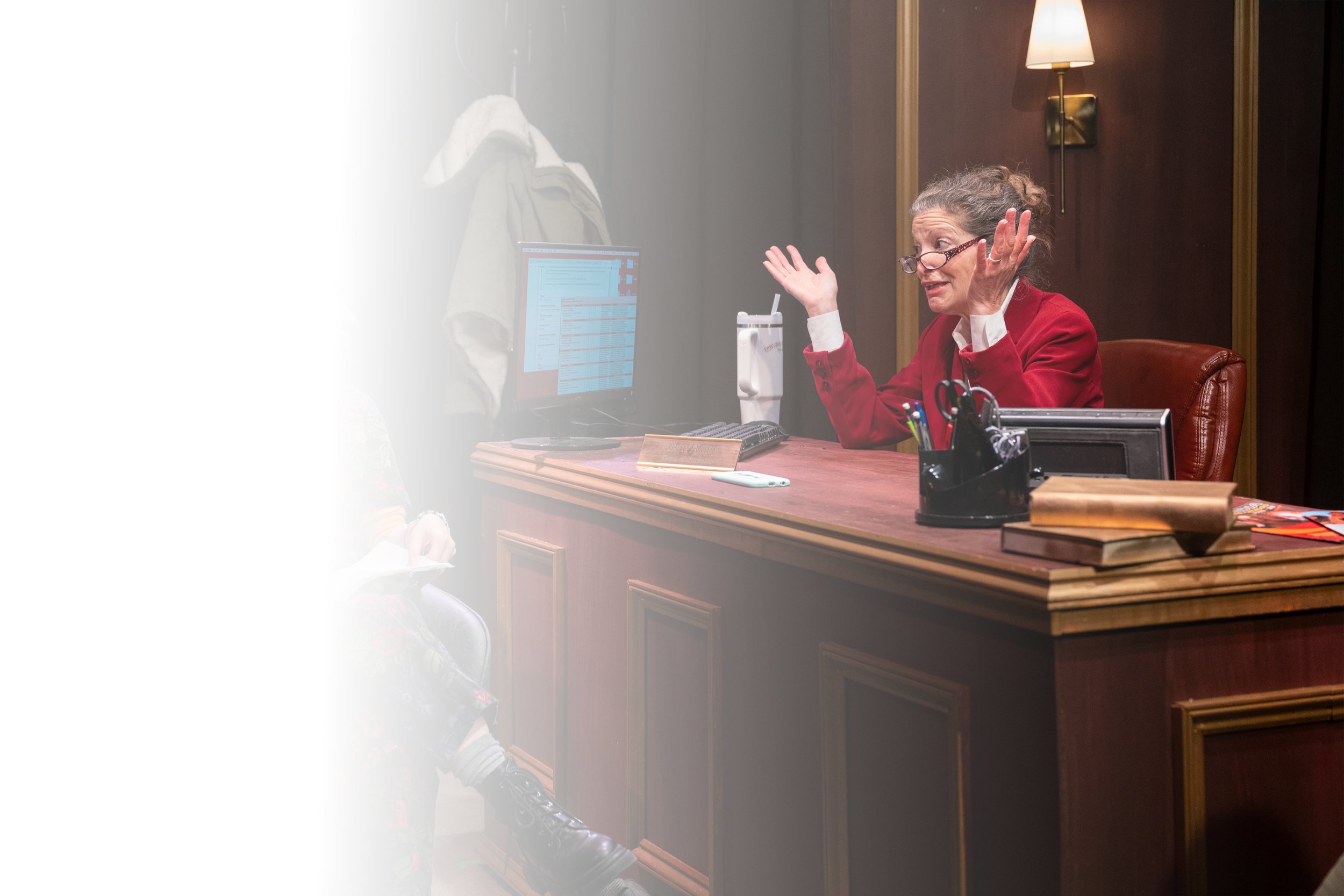 A woman with glasses and gray hair in a red blazer and white shirt talking to a person sitting on a black chair, wearing a colorful floral patterned outfit. The woman is gesturing with her hands, and the person is holding papers. They are in an office with a wooden desk, a computer monitor, books, a pen holder, and a desk phone. There is a coat rack with jackets behind the woman and a wall lamp providing warm lighting.