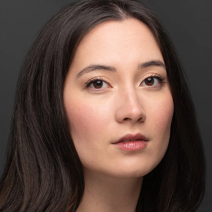 Close-up portrait of a young woman with long dark brown hair, fair skin, and brown eyes, looking slightly to the side against a dark background.