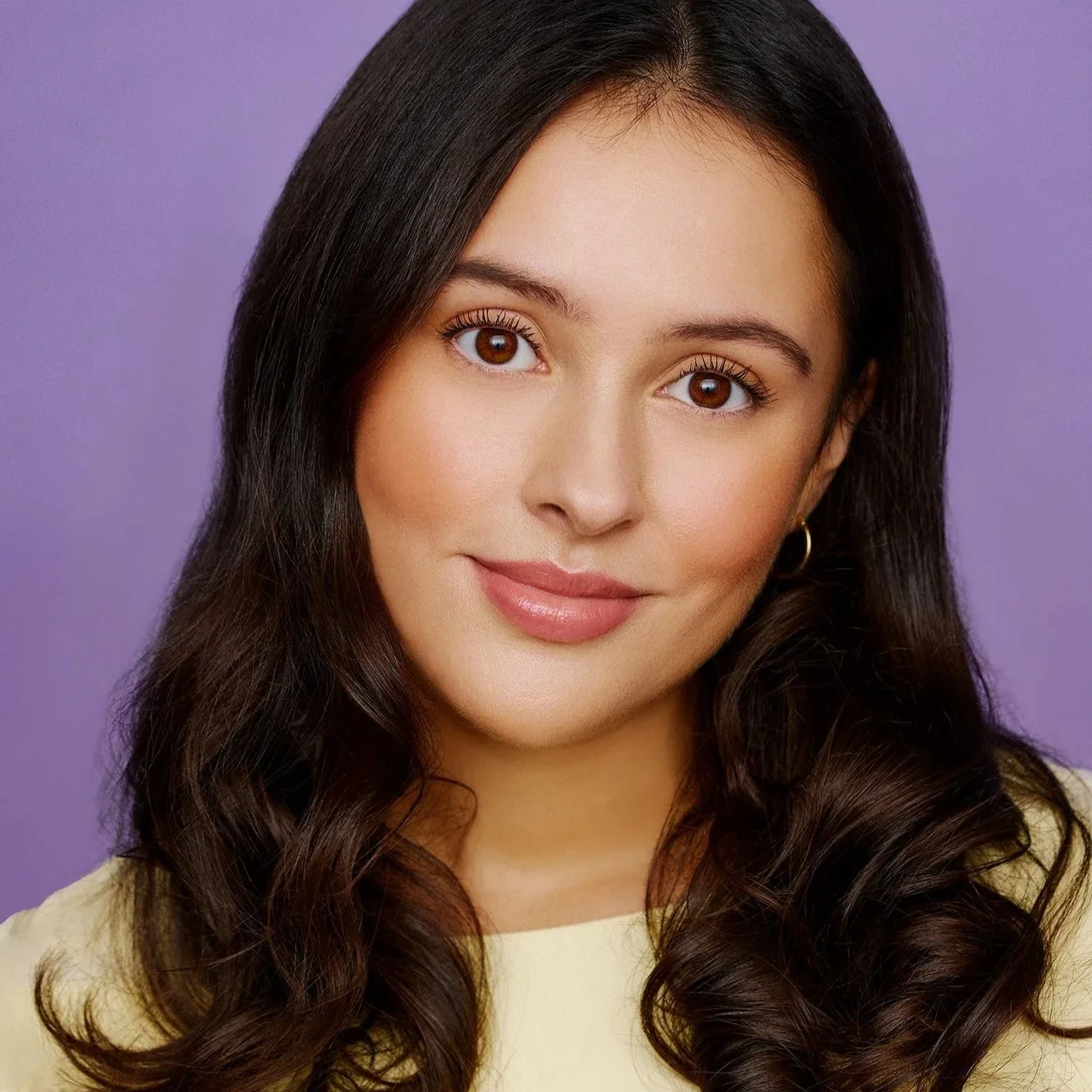 A young woman with long, dark, wavy hair wearing a light-colored top and small hoop earrings, smiling softly against a purple background.