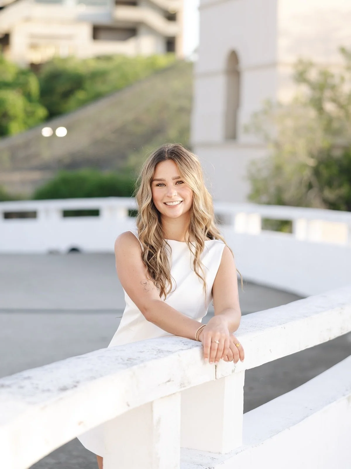 A smiling woman with long wavy blonde hair, wearing a white sleeveless dress, leaning on a white railing outdoors with a white building and greenery in the background.