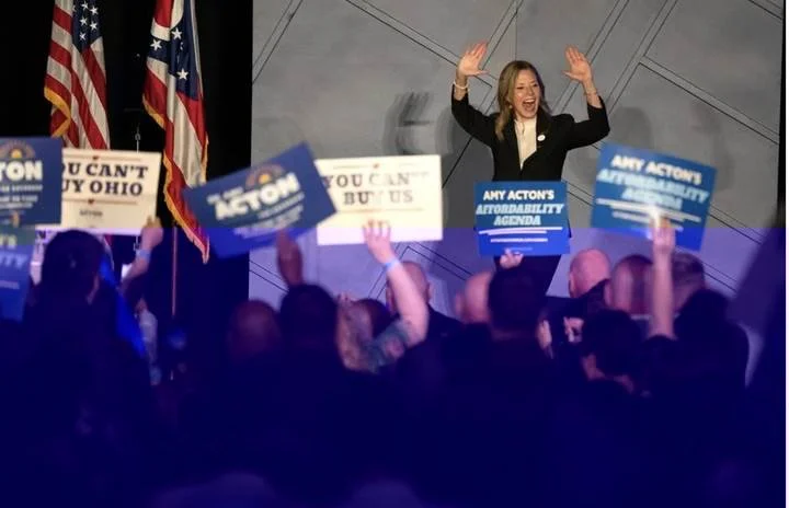 Amy Acton on stage at a campaign event, arms raised, supporters holding signs