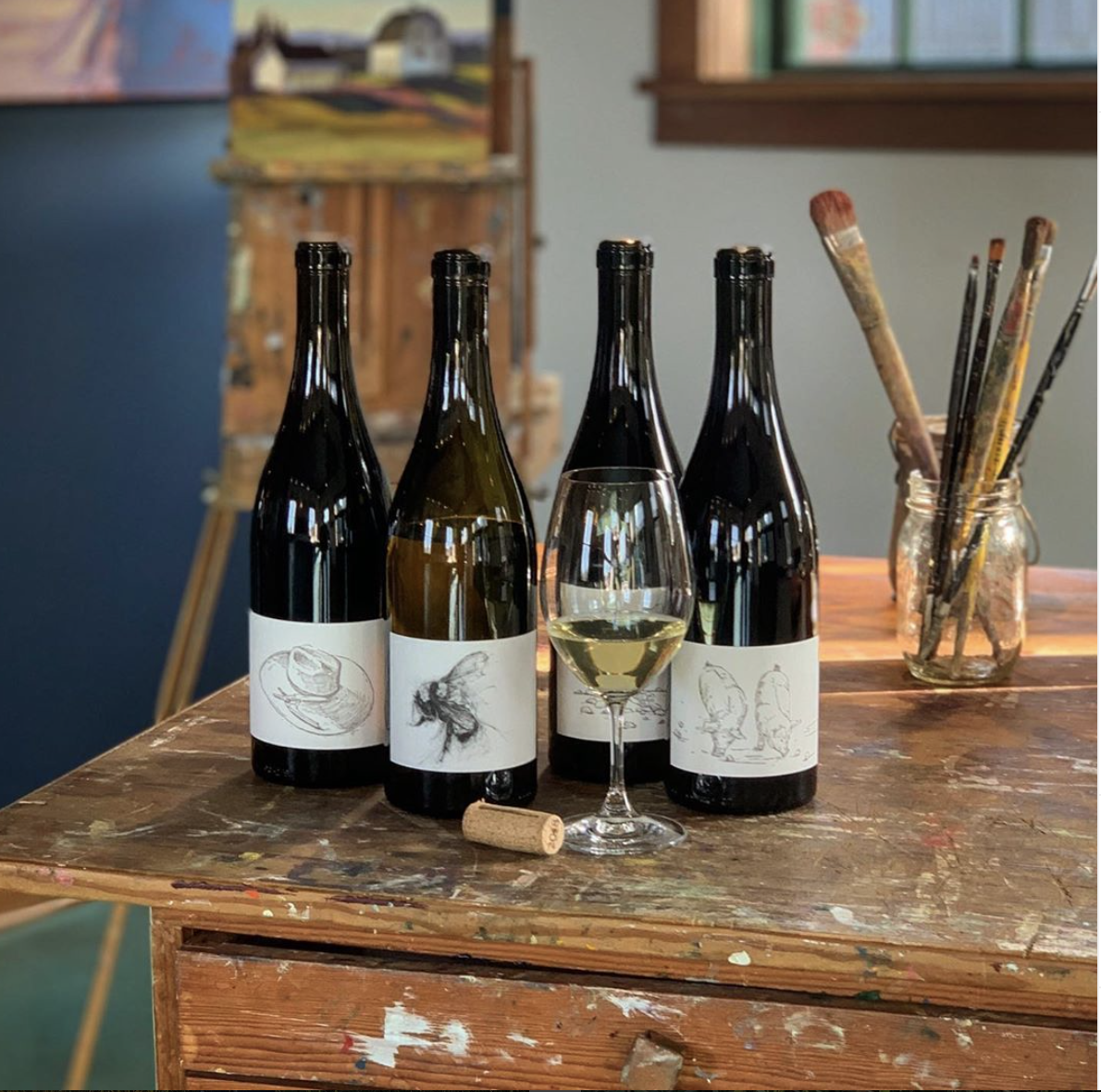  Four bottles and a glass of Big Table Farm wine on a paint splattered art table in the Atelier in Carlton, OR. 