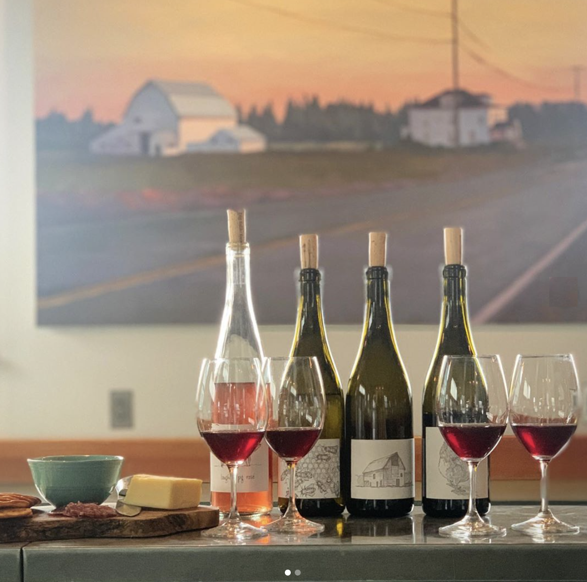  A wine tasting at the Big Table Farm Atelier in Carlton, OR. Wine bottles, glasses, and cheese plate on a table with a  farm-scape painting by Clare Carver in the background. 