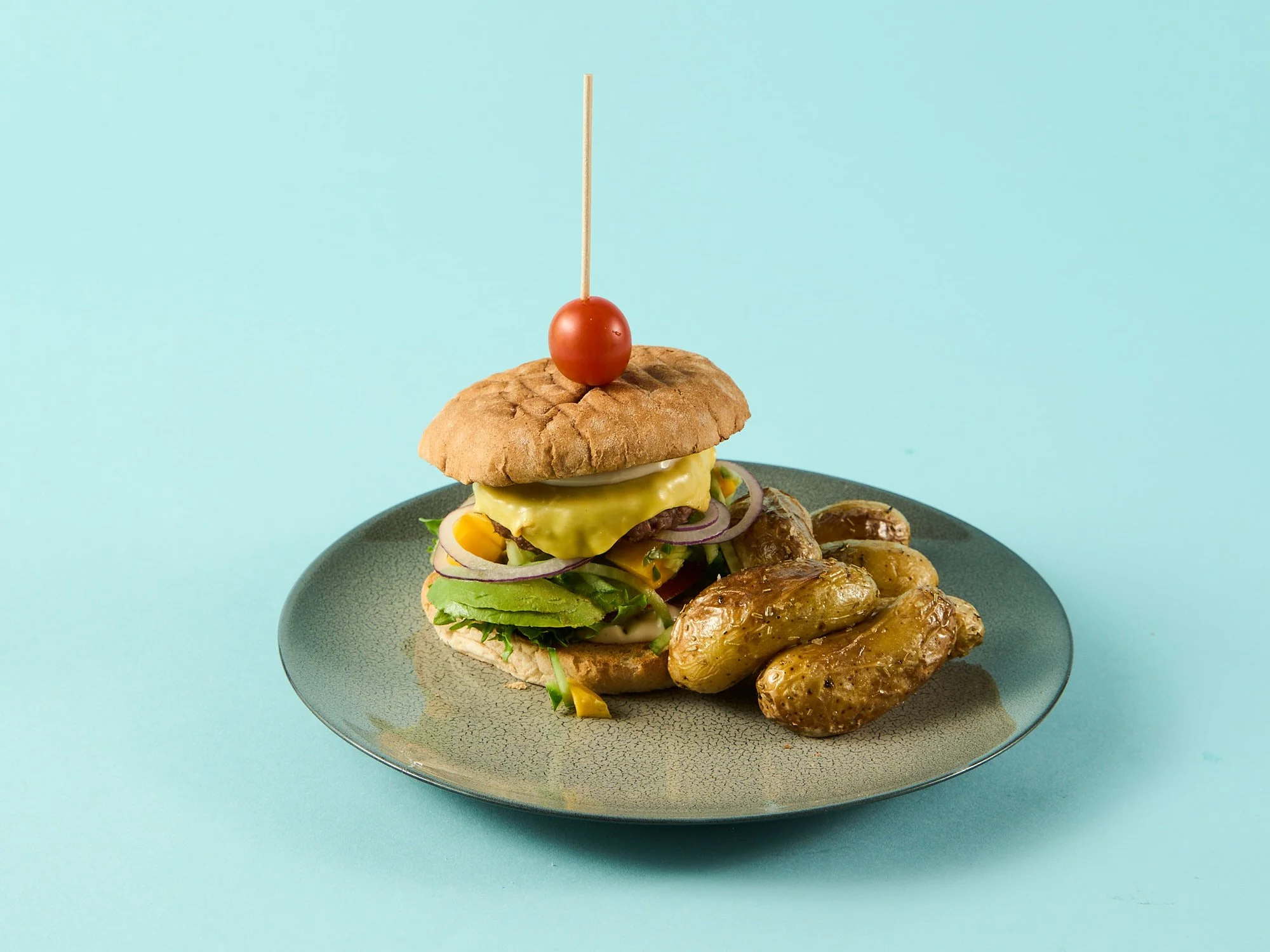 En burger med ingredienser som salat, løk, ost, kjøttdeig, og en liten tomat på en treplate med poteter ved siden av, mot en blå bakgrunn.