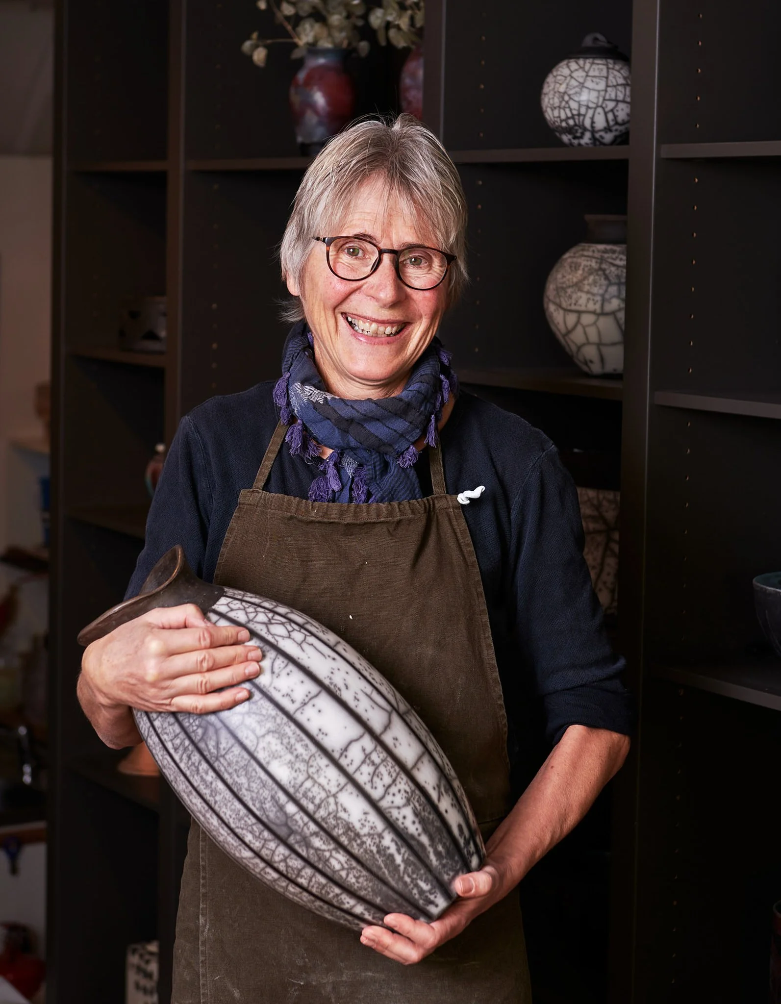 En eldre kvinne med kort grått hår, briller og smilende ansikt, holder en keramisk sculpture med svart og hvitt mønster, står foran en bokhylle med dekorative vaser.