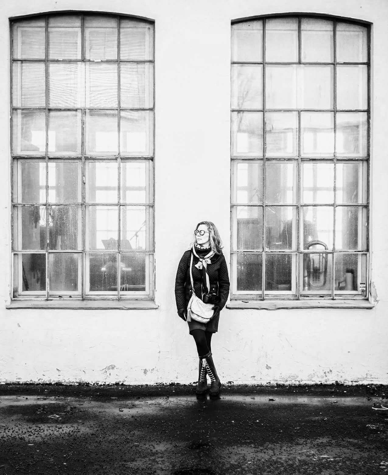 En kvinne med briller står foran to store vinduer på en hvit vegg, iført svart kåpe og bukse, med veske og sko med hæler. Fotografiet er i svart-hvitt.