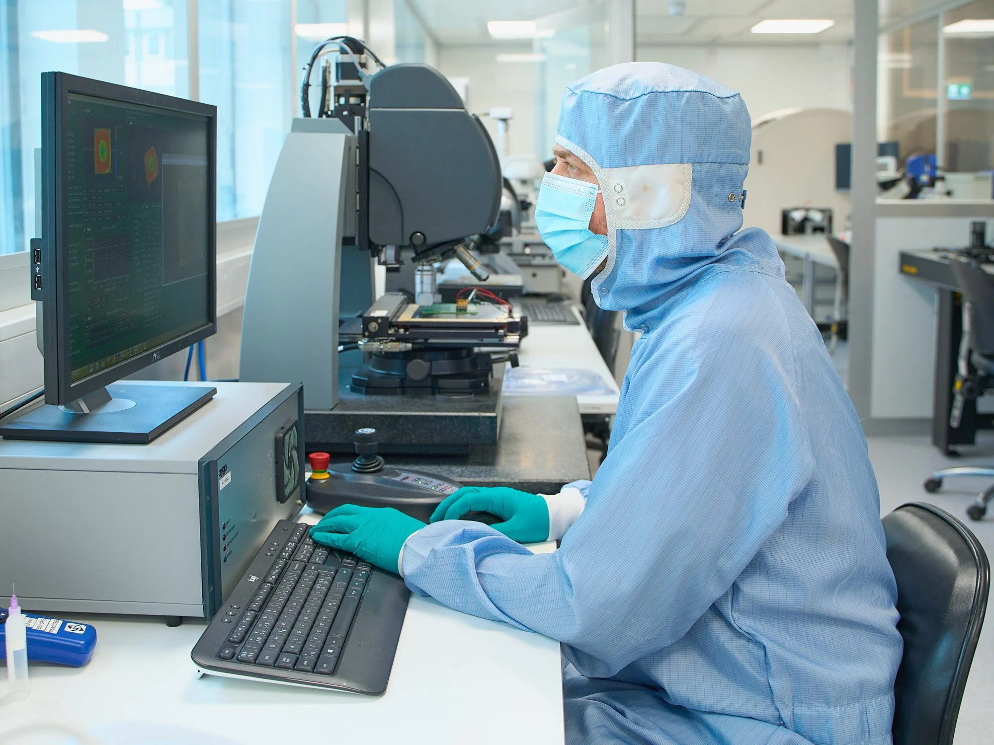 Person i beskyttelsesdrakt og maske som jobber med laboratorieutstyr foran en dataskjerm i et moderne laboratorium.
