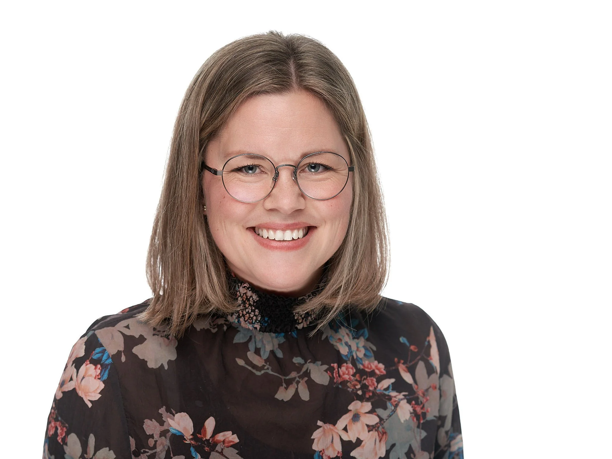 Et smilende kvinnes ansikt med brunt langt hår, iført briller og en blomstrete topp.
