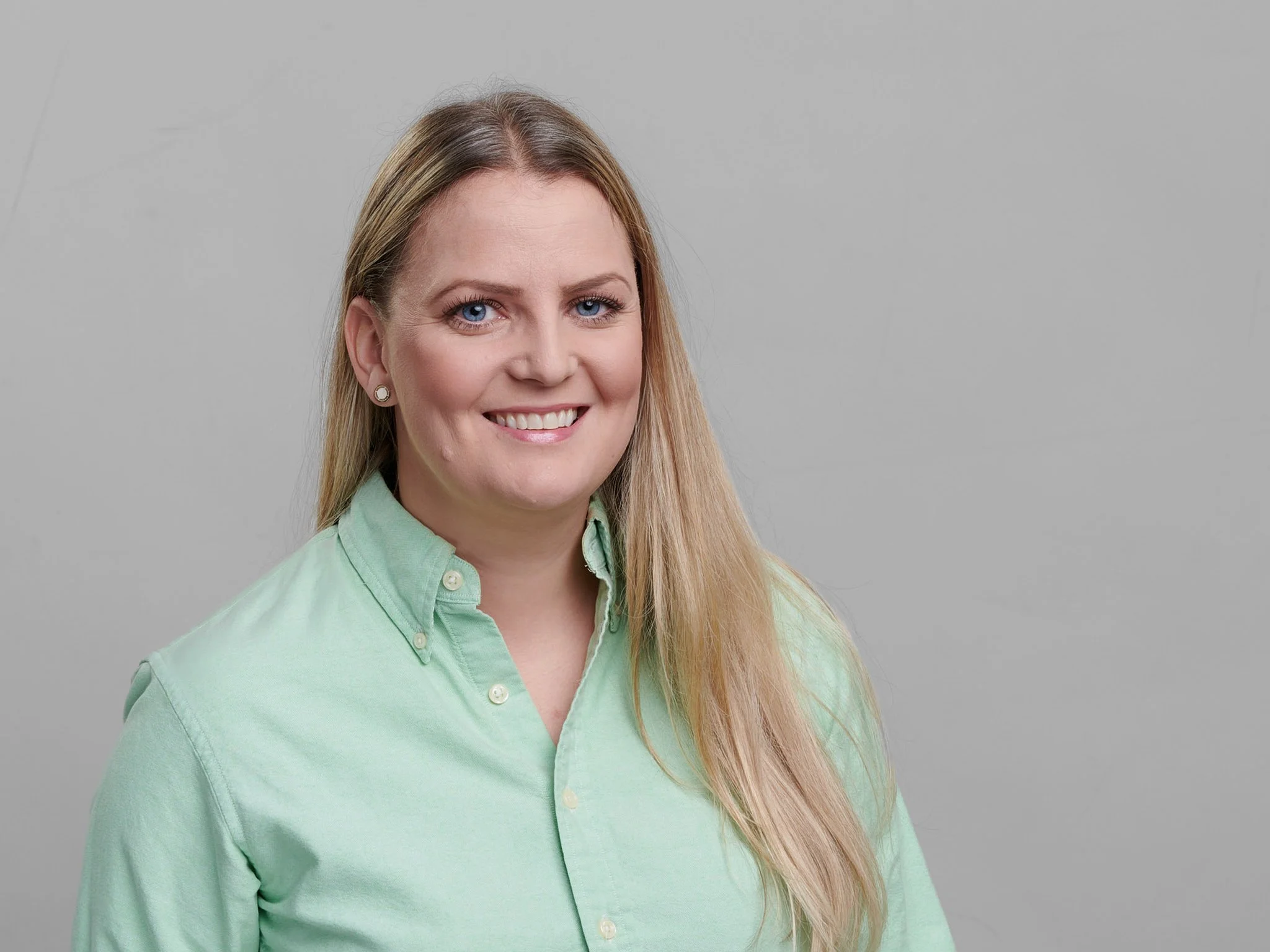 Et bilde av en smilende kvinne med lang blond hår, iført en lys grønn knappet skjorte, stående foran en grå bakgrunn.