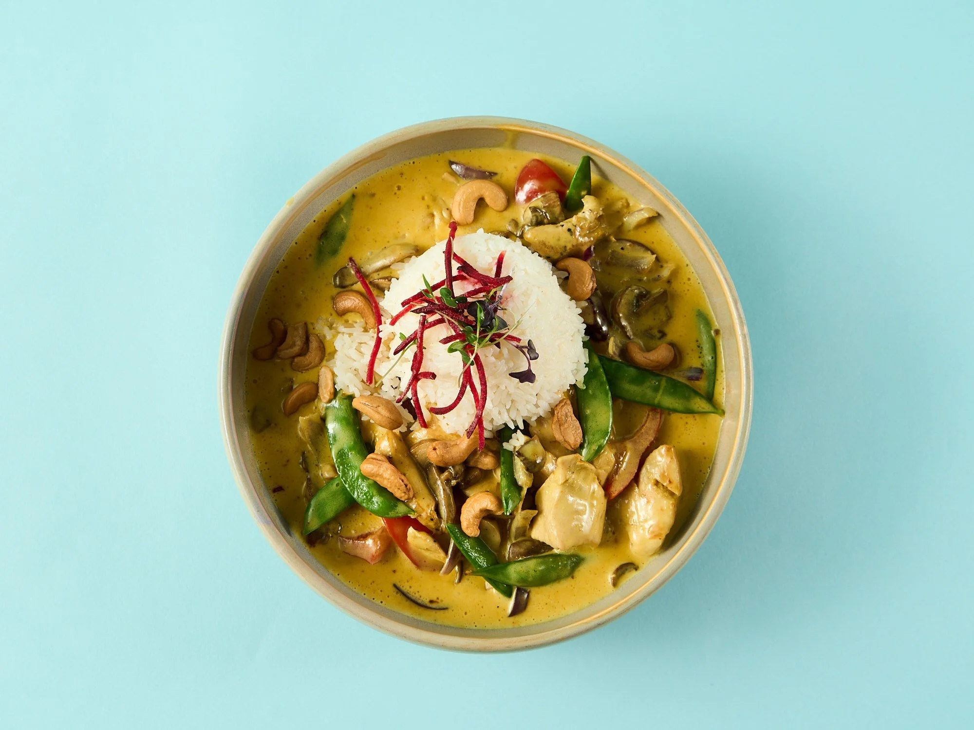 Bilde av en bolle med grønnsaks-curry, servert med ris, pyntet med urter og cashewnøtter, mot en lys blå bakgrunn.