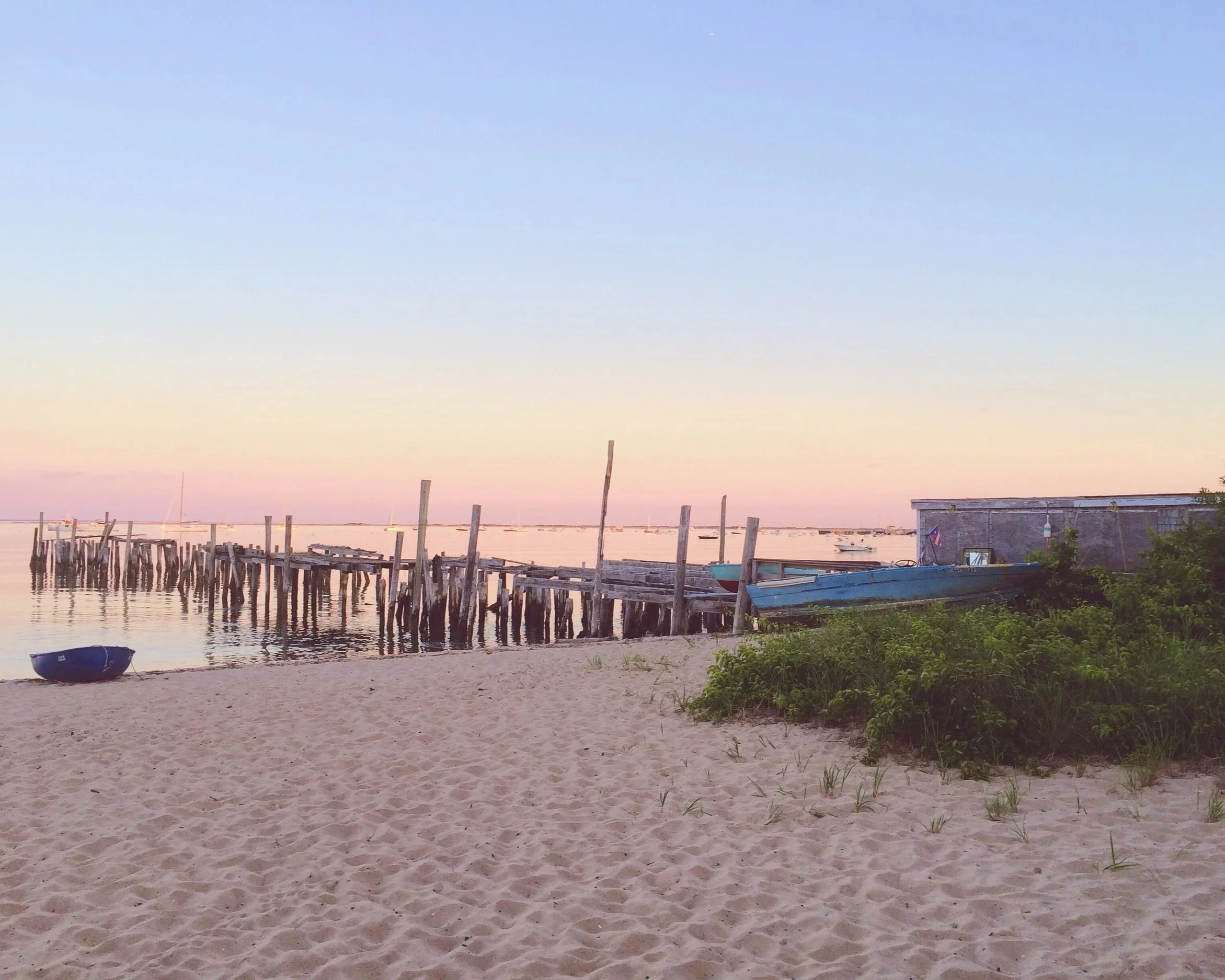 A sandy beach with a small blue boat on the shore, a weathered wooden pier extending into the water, and a rustic building with a boat nearby, under a pastel-colored sky at sunset.
