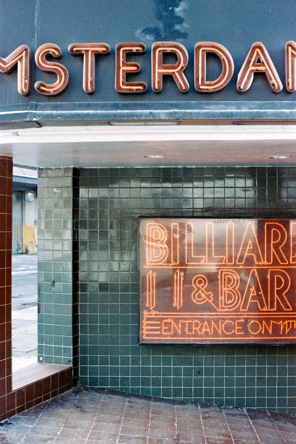 Neon sign reads 'AMSTERDAM' and a smaller sign reads 'BILLIARDS 11 & BAR ENTRANCE ON THE' with the text partially cut off, displayed in green tiled exterior wall.
