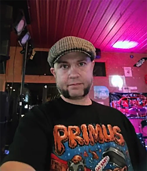 A man wearing a brown checkered flat cap and a black graphic T-shirt standing in a room with red wooden walls, neon lights, and musical equipment.