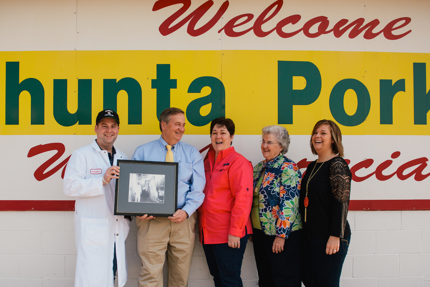 Bradon, Larry, Rita, Jean, and Lauren pose for a photo with a portrait of Mack "PigPop" Pierce holding his largest and most prized 1,105 pound salughtered hog. The Pierce family runs the Nahunta Pork Center and Hog Market, keeping family values and respect at the core of their business.