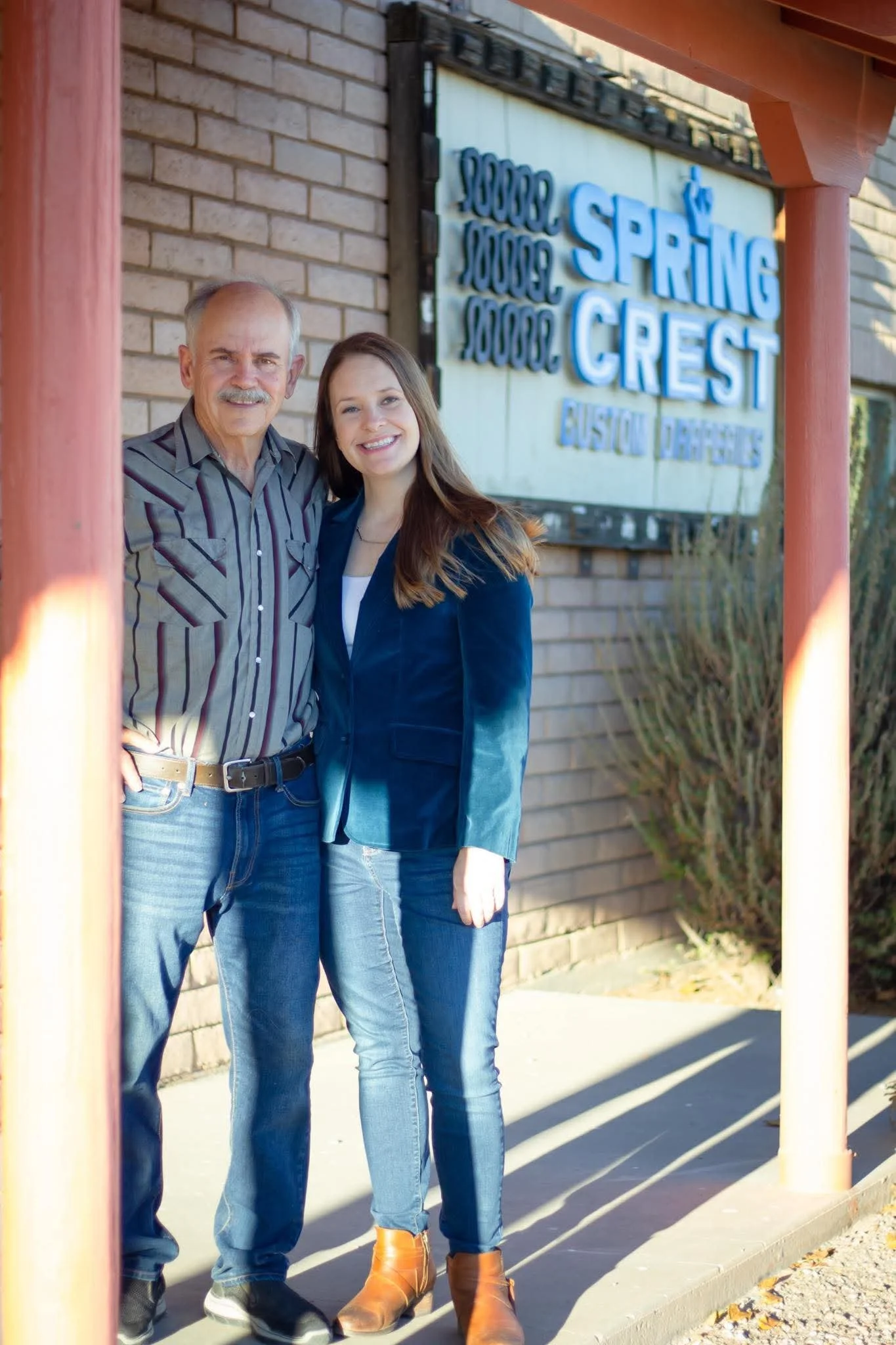 A man and a woman standing outside a business called Spring Crest, smiling for a photo. They are positioned under a red wooden frame, with the business sign visible behind them. The man has gray hair, a mustache, and is wearing a striped button-up shirt and jeans. The woman has long brown hair, and is wearing a blue blazer, white shirt, jeans, and tan boots.