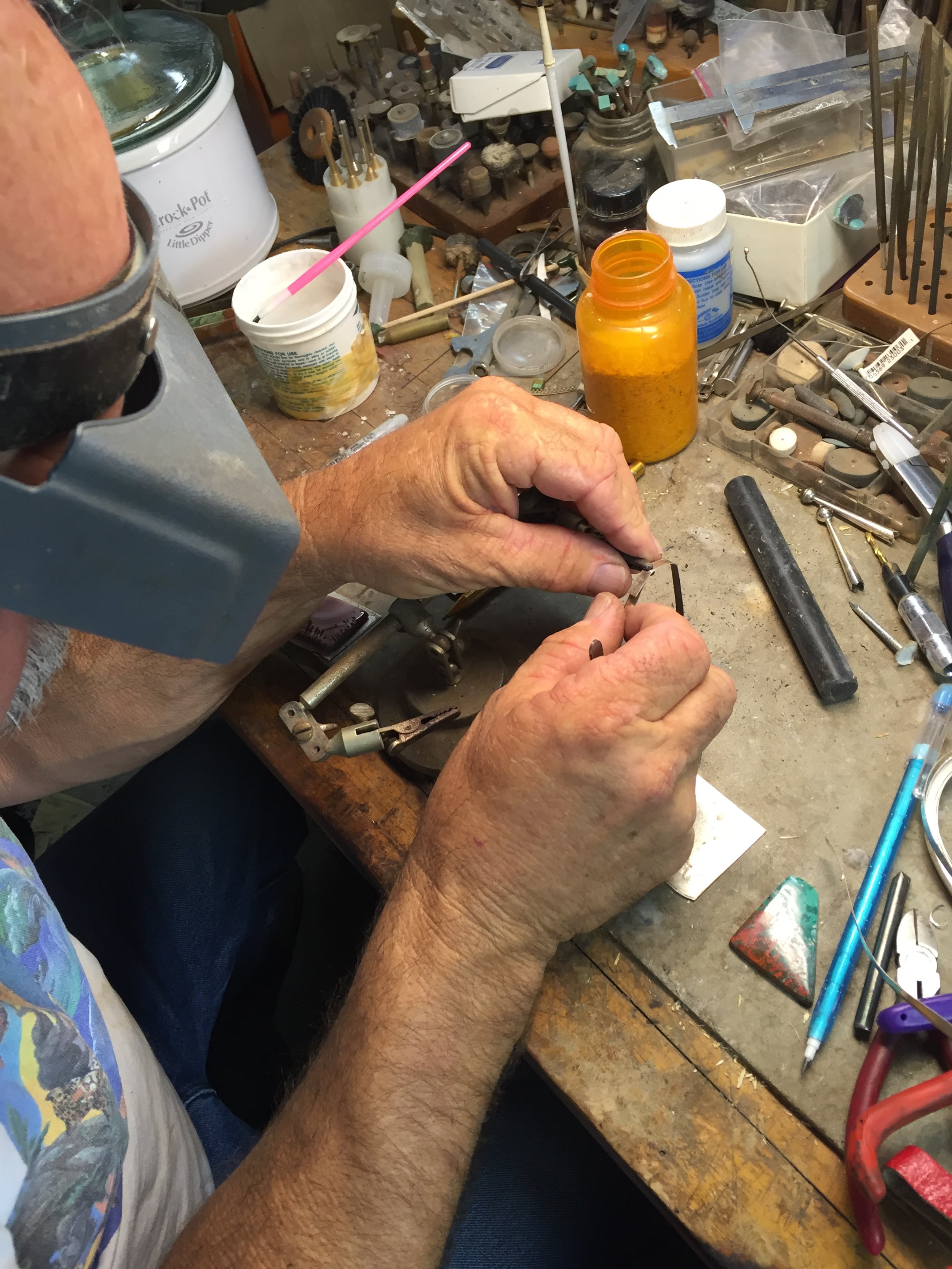 Close-up of elderly man wearing magnifying glasses, working on jewelry at cluttered workbench with tools, small containers, and supplies.