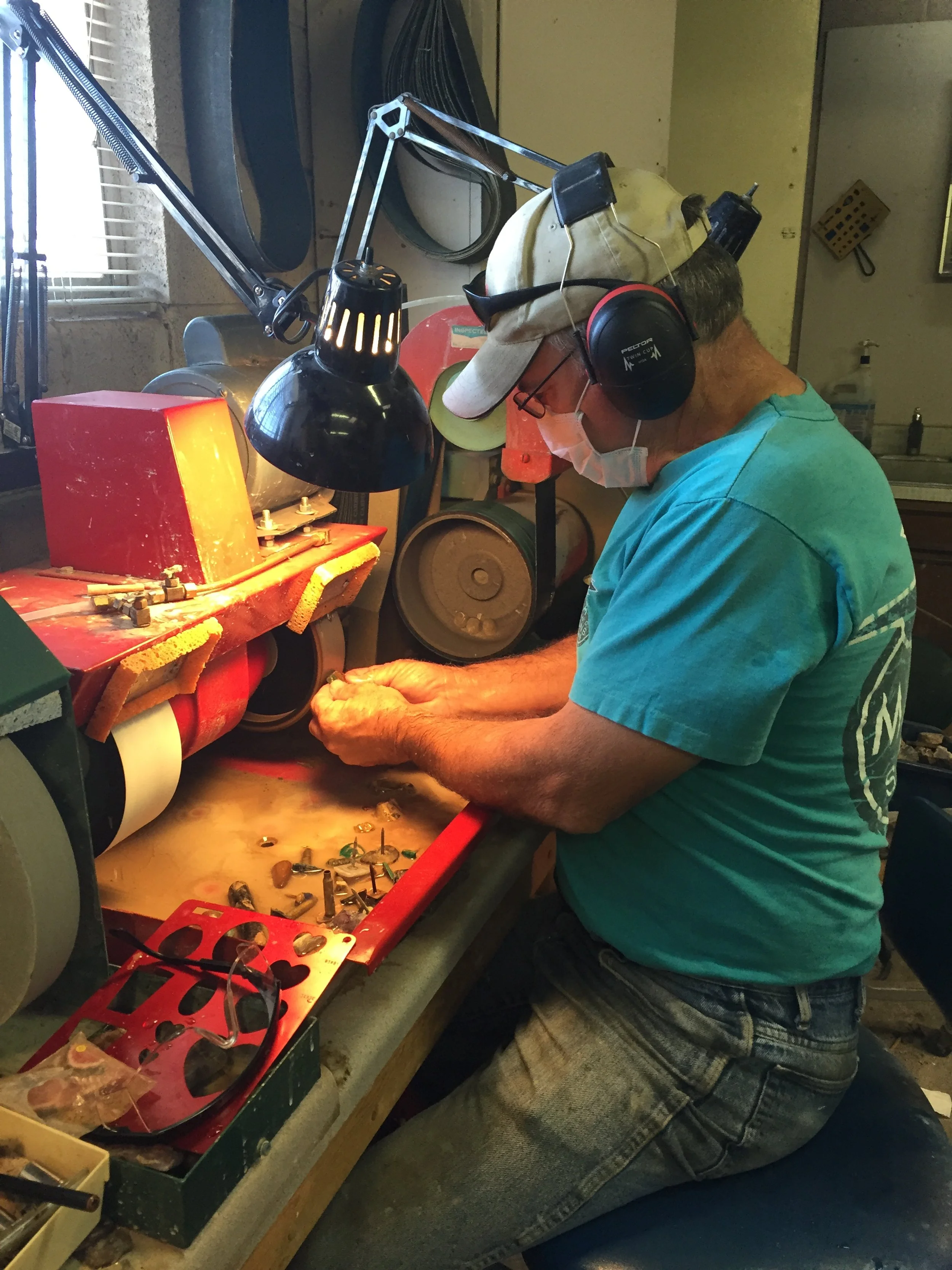 A man wearing glasses, a mask, headphones, and a cap is working at a jewelry polishing or finishing station. He is using a handheld tool on a piece of jewelry, with various tools and equipment around him on the workbench.