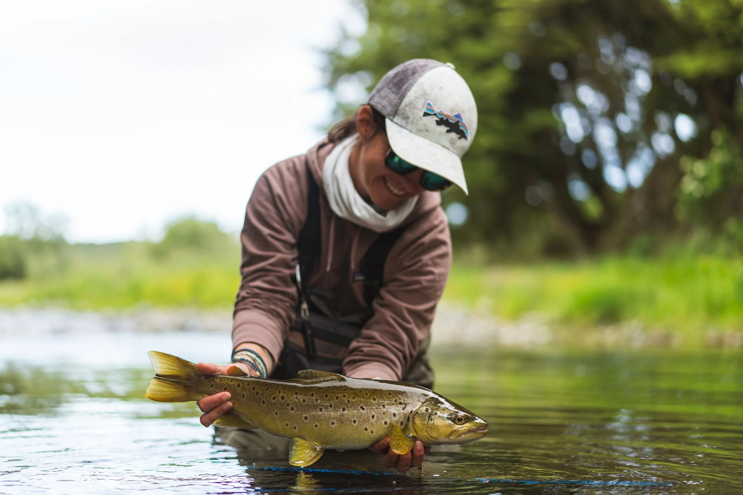 A person wearing a baseball cap, sunglasses, and outdoor clothing holding a large fish in shallow water, with trees and a bright sky in the background.
