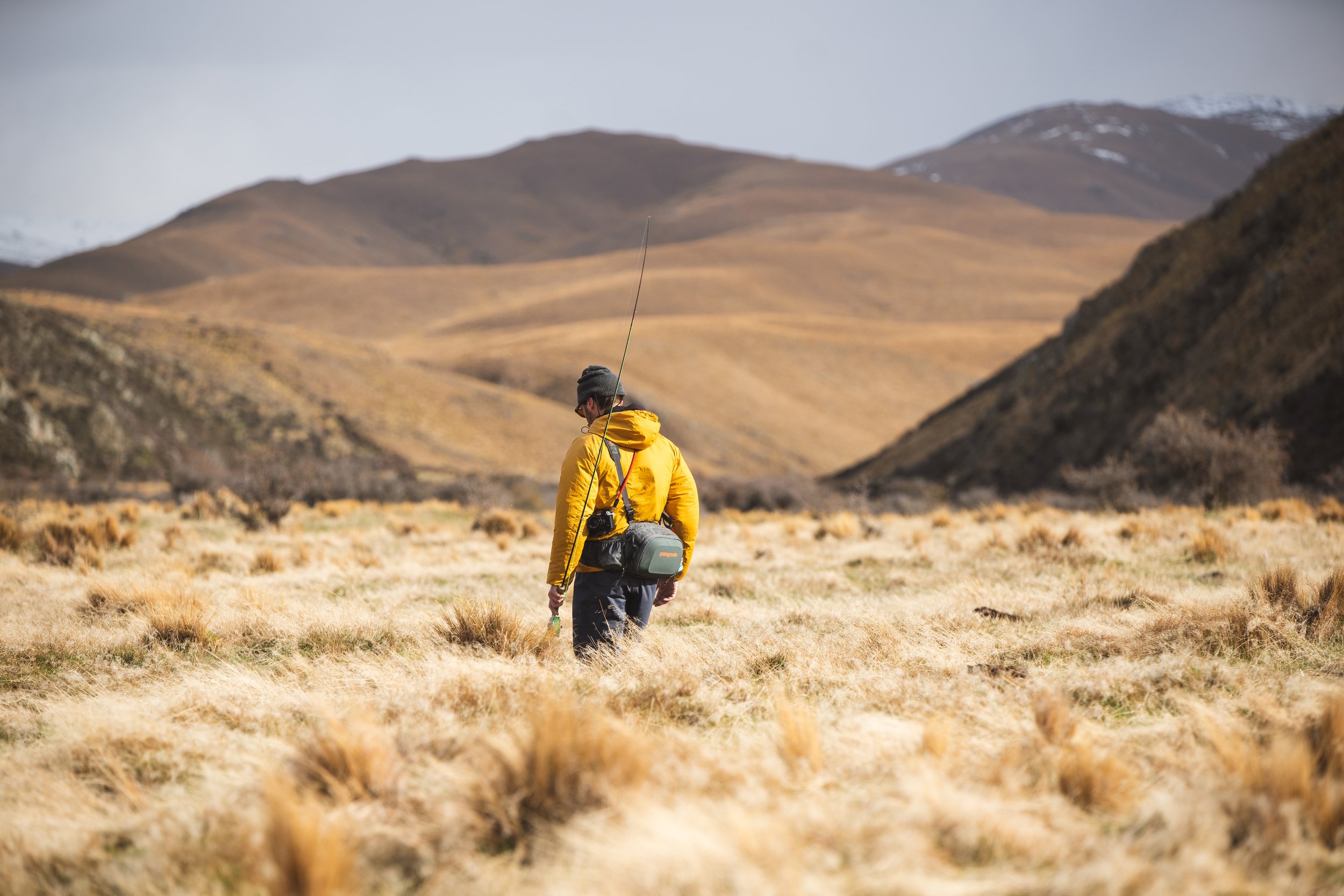 A person in a yellow jacket and gray hat with a camera on their waist walks through a grassy field in a mountainous area, with brown hills and snow-capped mountains in the background.