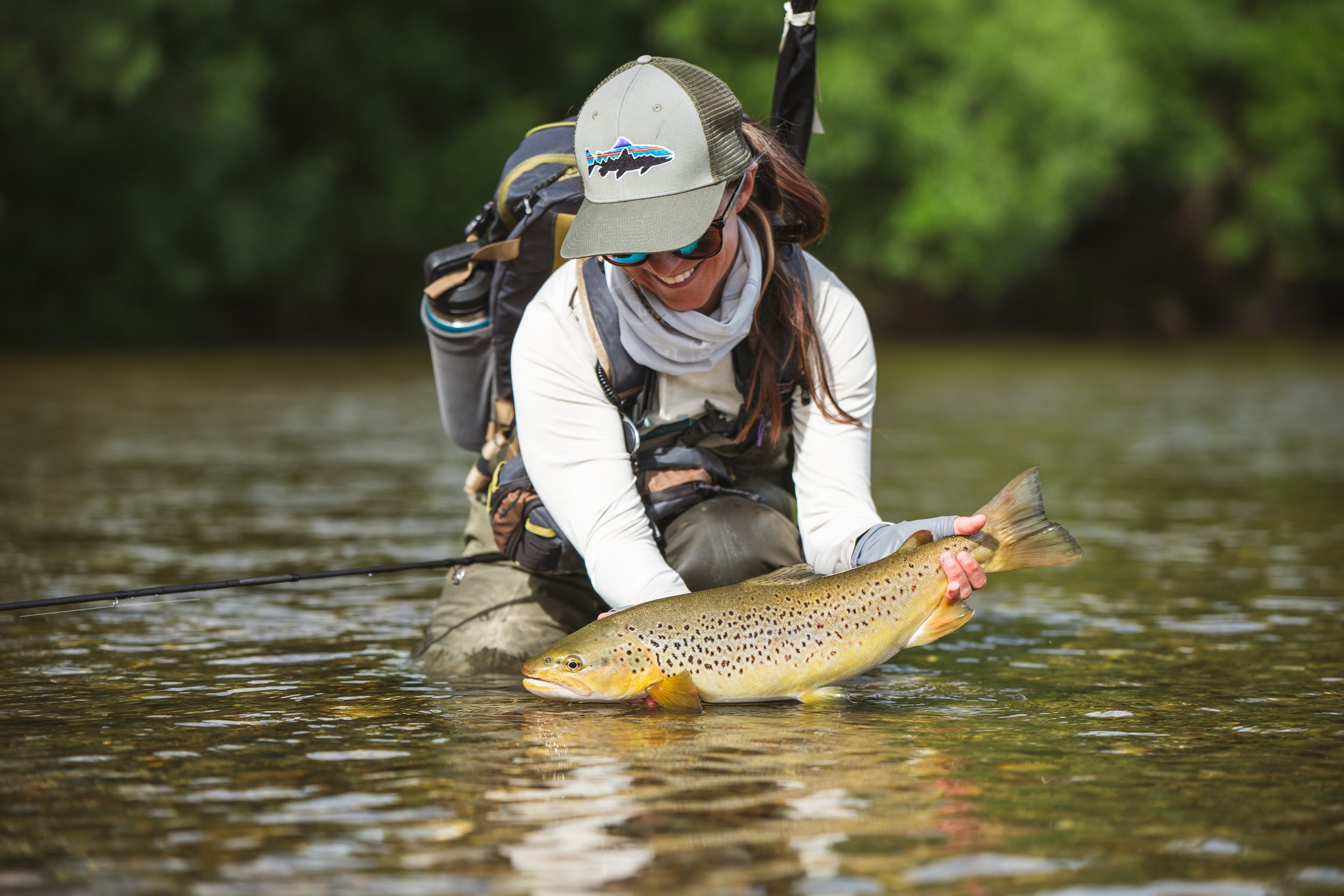 A woman in outdoor gear, wearing a cap and sunglasses, kneeling in a shallow river, holding a large brown trout she caught while fishing.
