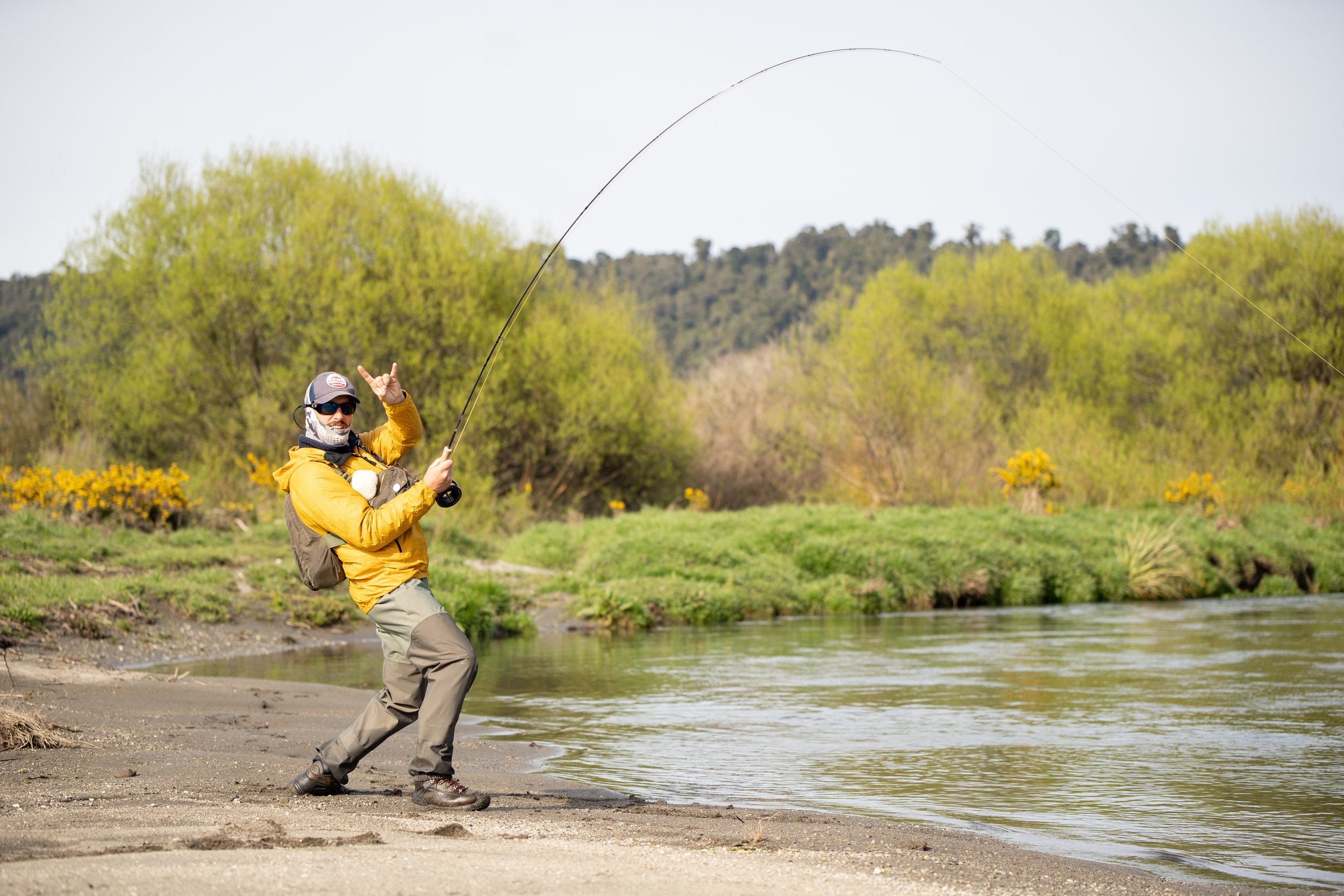 A person in outdoor fishing gear standing on a lakeshore, casting a fishing line into the water.