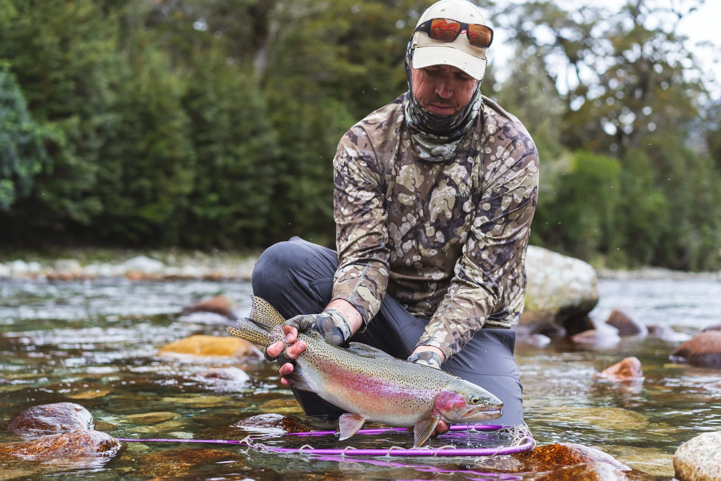 A man in camouflage clothing and a white cap holds a large fish over a shallow river, with rocks and trees in the background.