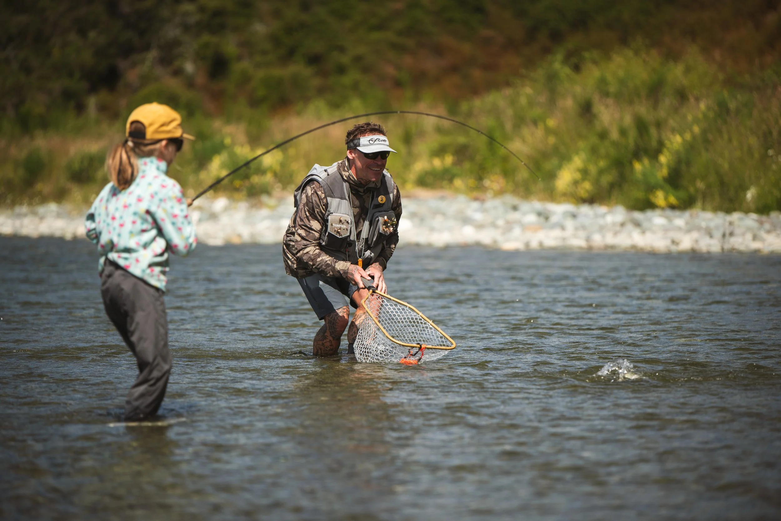 Two individuals fishing in a river with a lush, green backdrop. One person is a man wearing sunglasses, a sun visor, and fishing gear, holding a fishing net. The other person, a girl, is wearing a cap, sunglasses, and a colorful jacket, holding a fis