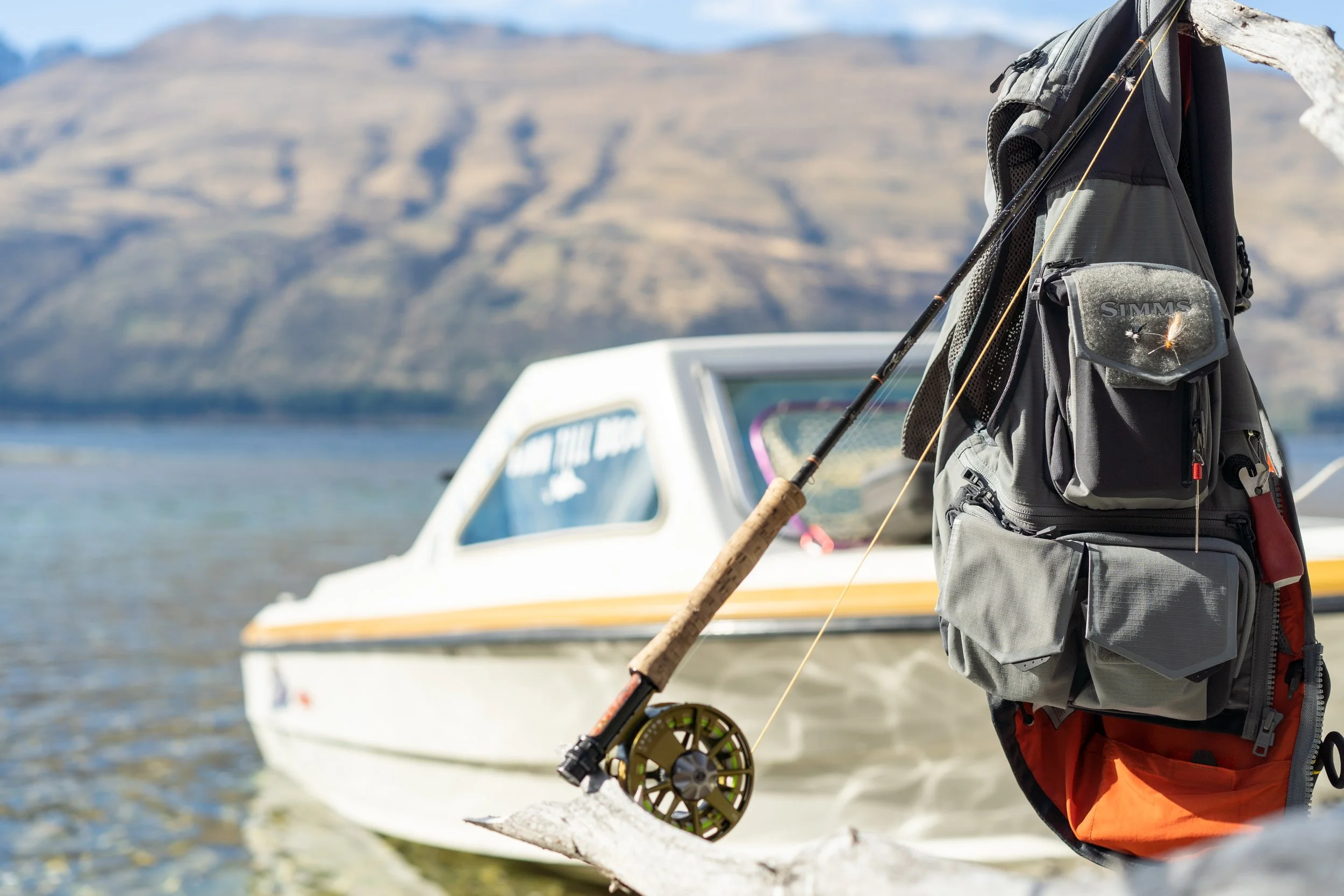 A person with a backpack and fishing gear standing on a beach, with a boat and mountains in the background.
