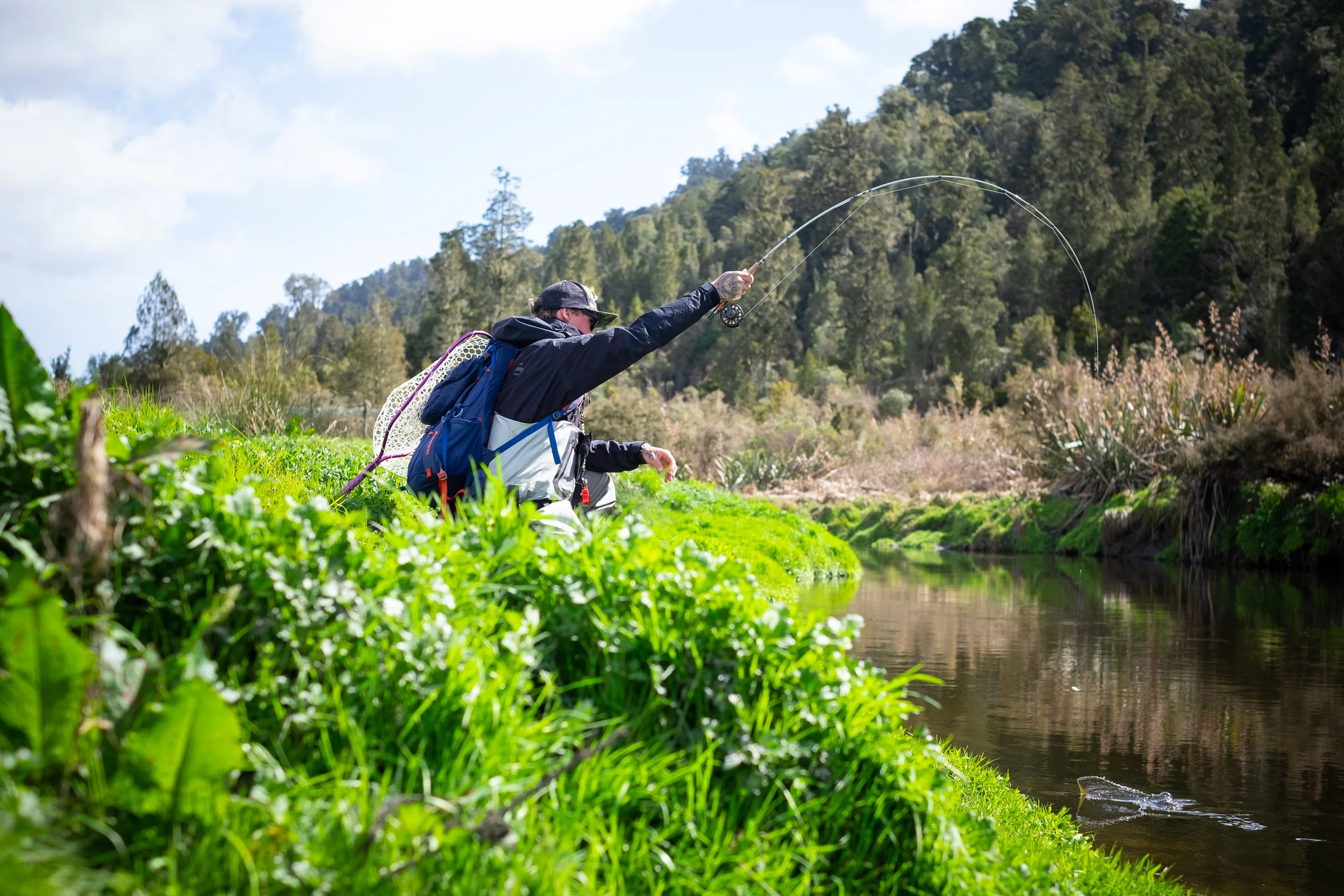 Person fishing by a stream in a lush green landscape with trees and mountains in the background.